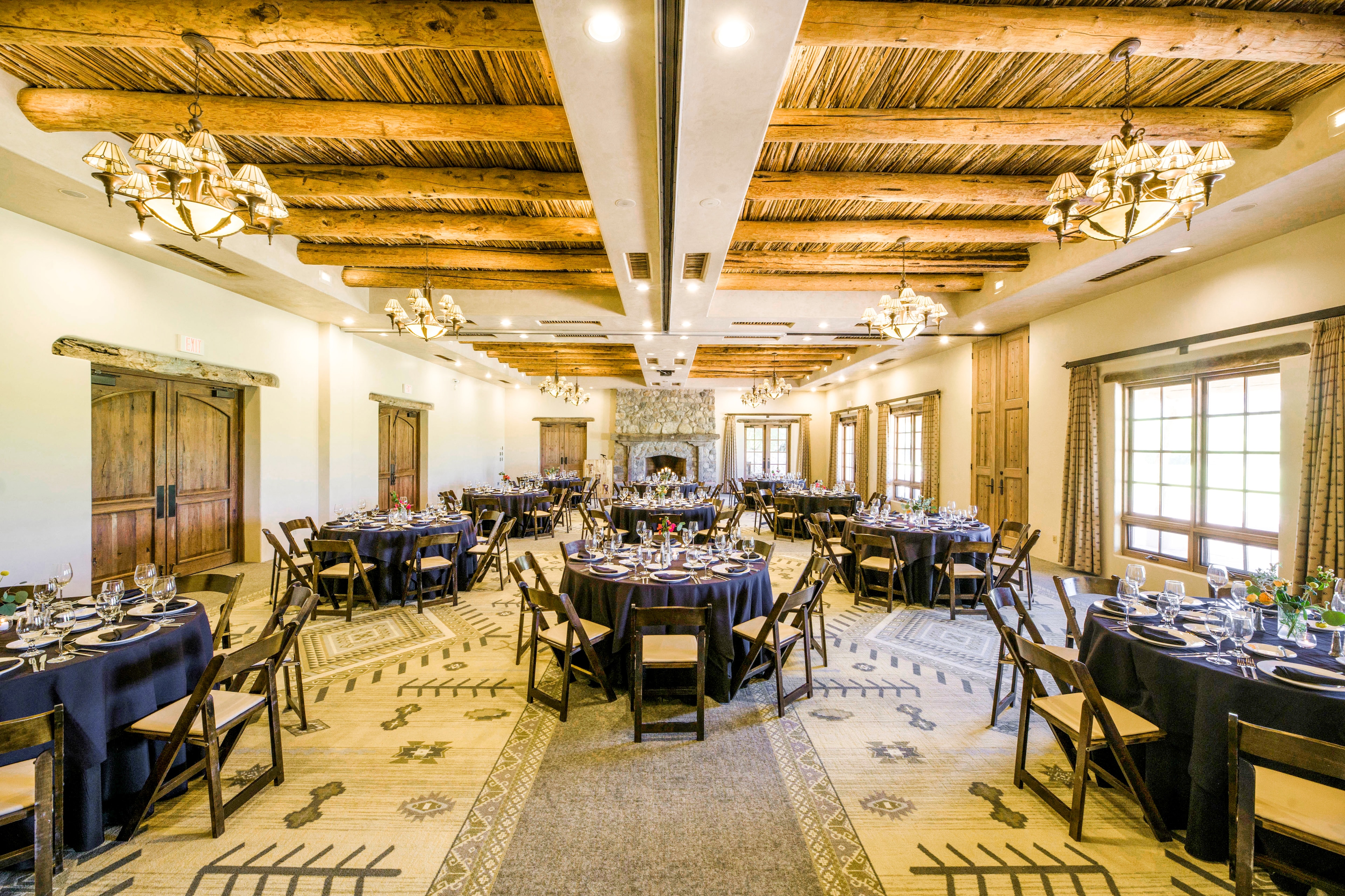 A large dining room with round tables, wooden chairs, exposed beams, chandeliers, and a fireplace.