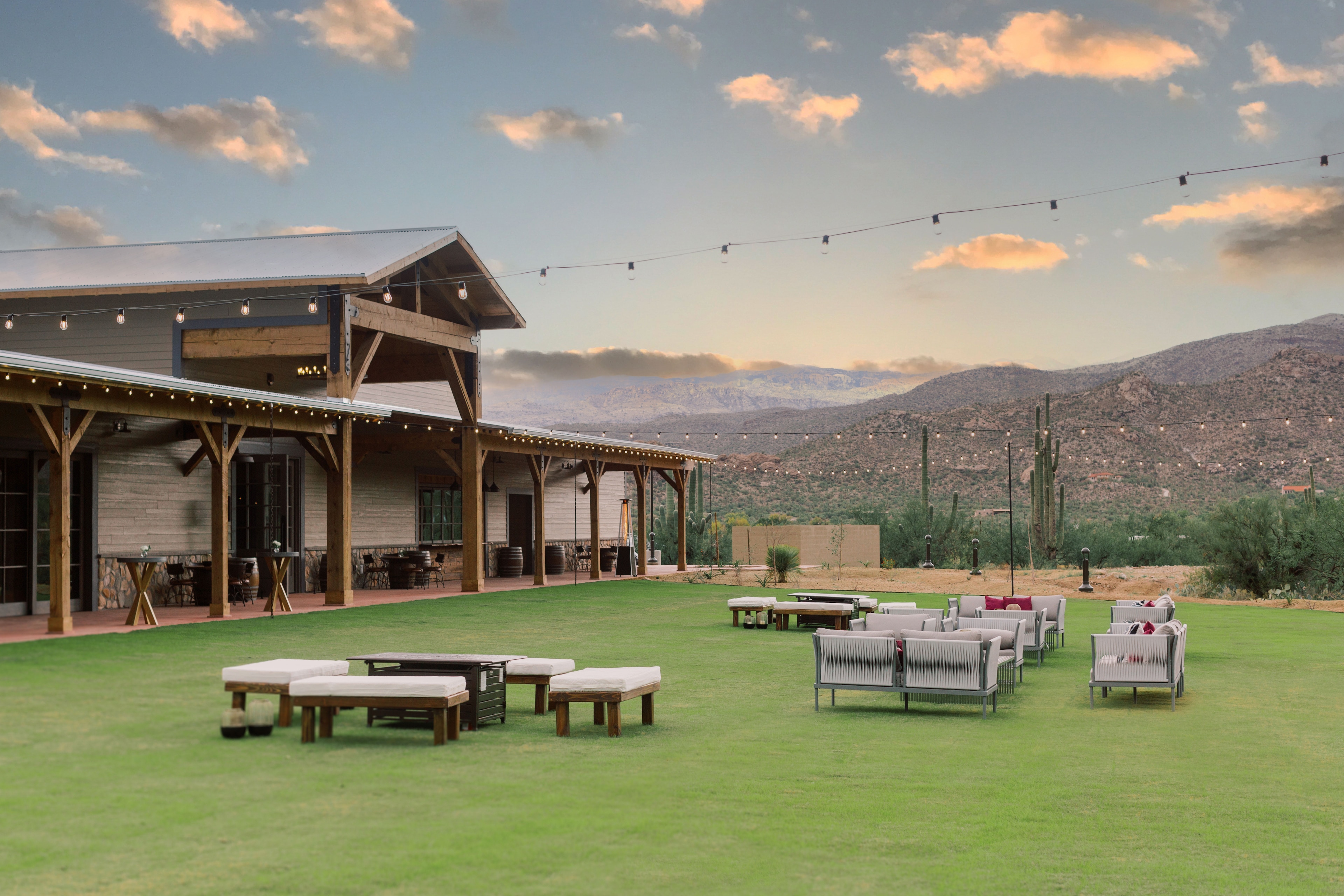 A large building with a wooden structure and string lights, surrounded by a grass lawn and outdoor seating areas, with a mountain view in the background.