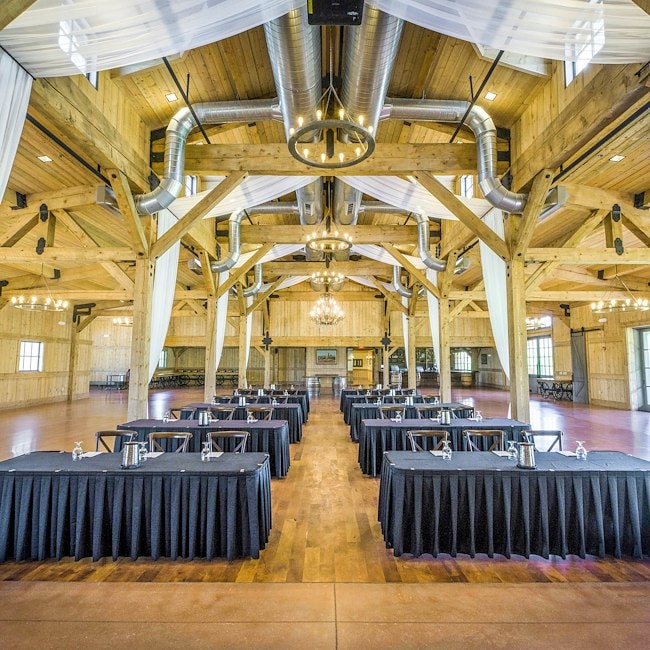 A large, rustic meeting room with wooden beams, exposed ductwork, and long tables with black tablecloths.