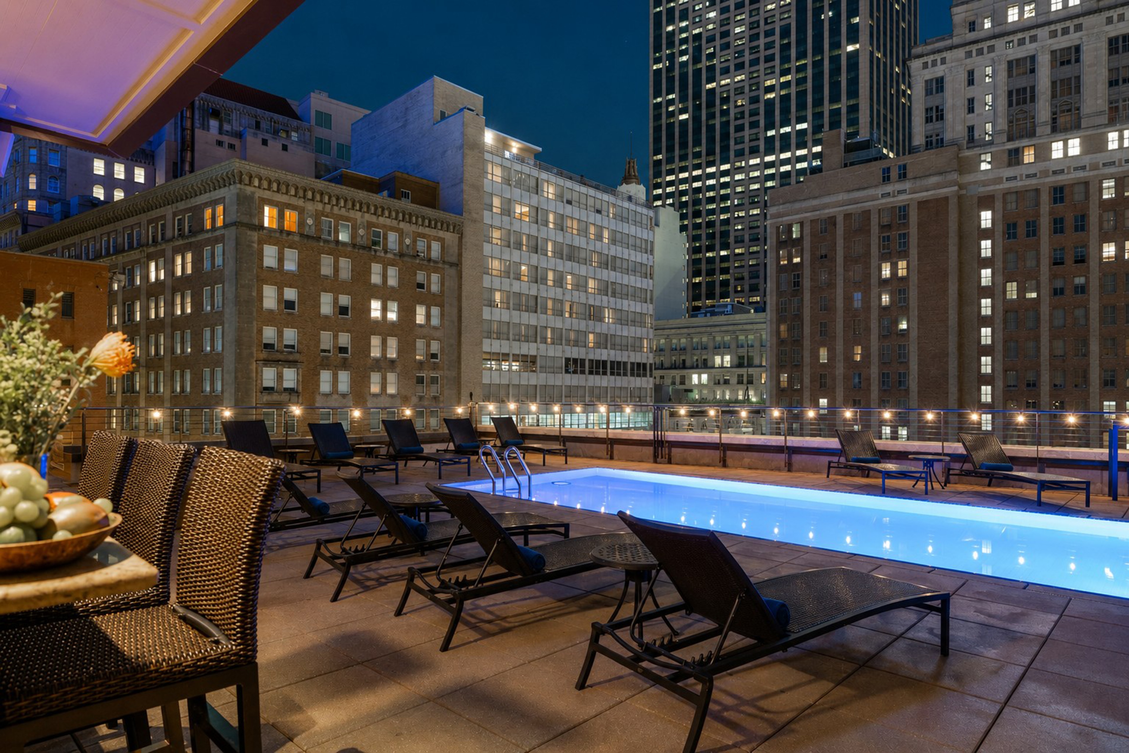 A rooftop pool with blue water and lounge chairs, overlooking a cityscape at night.