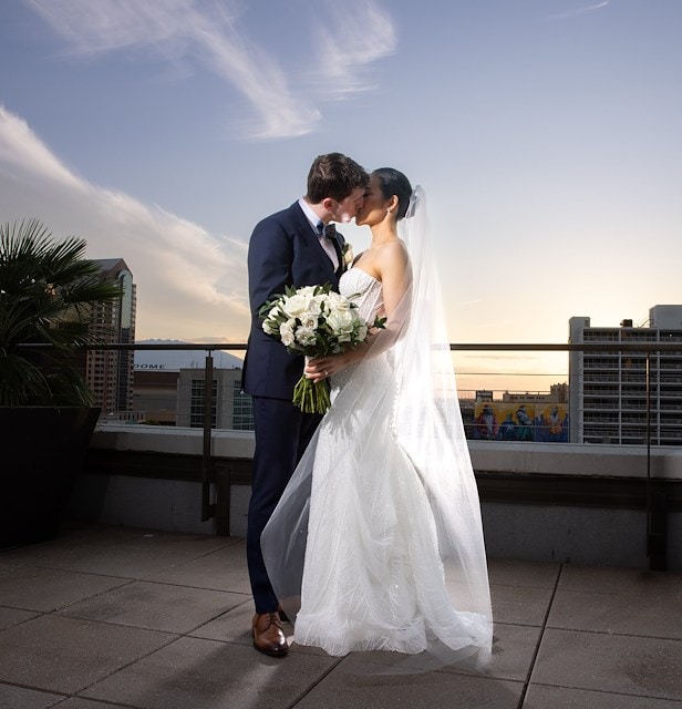 A couple in wedding attire on a rooftop with a city view at sunset.