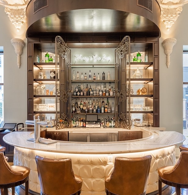A well-lit bar with a marble counter, leather chairs, and shelves stocked with bottles and glasses.