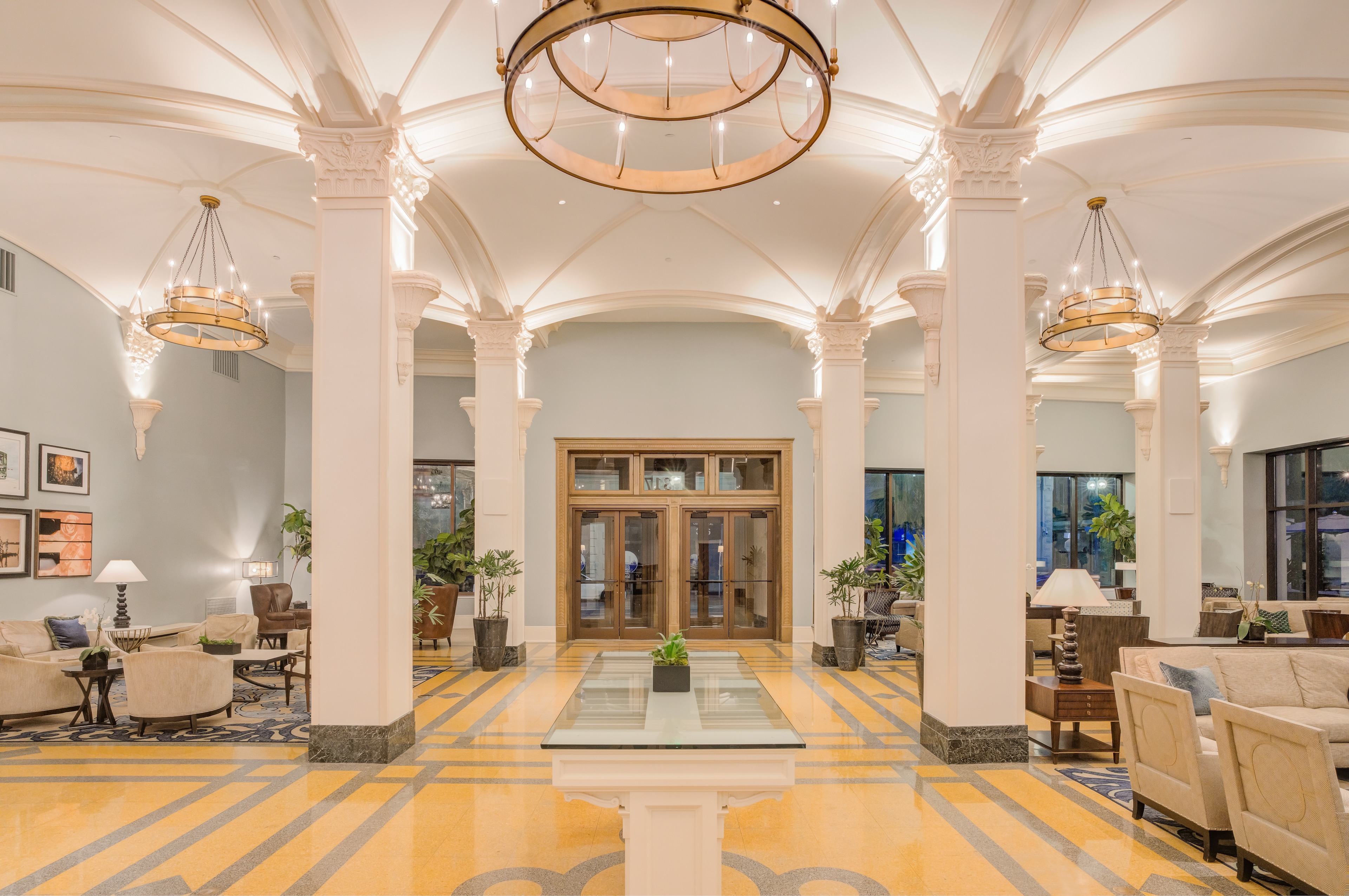 A wide view of a hotel lobby with a high ceiling, columns, chandeliers, and a glass table.