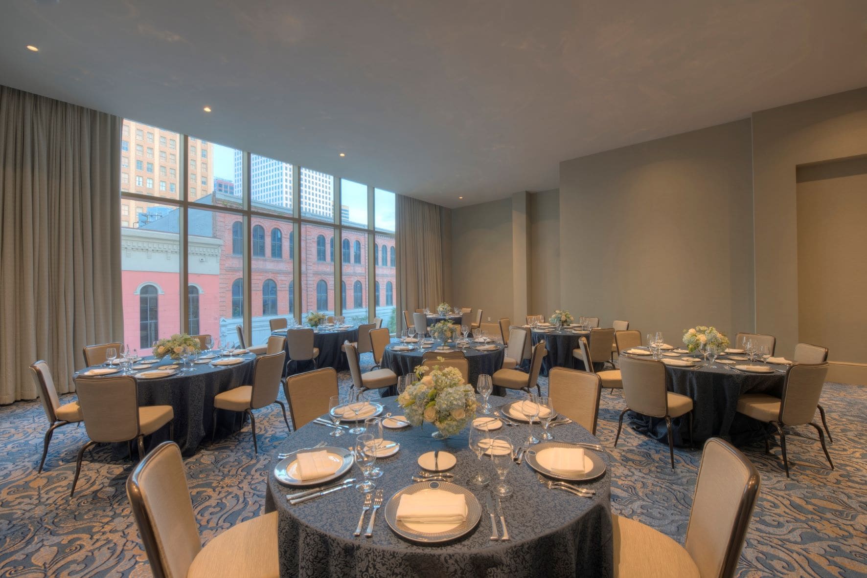 A formal dining room with round tables set for a meal, large windows, and a city view.