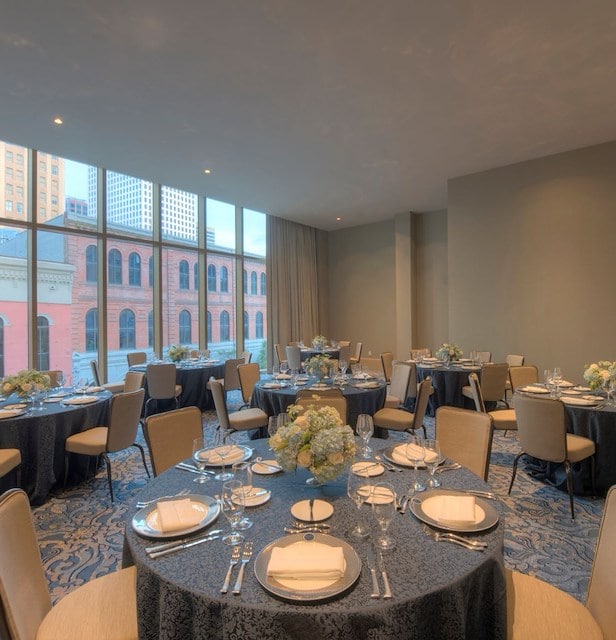 A formal dining room with round tables set for a meal, large windows, and a city view.