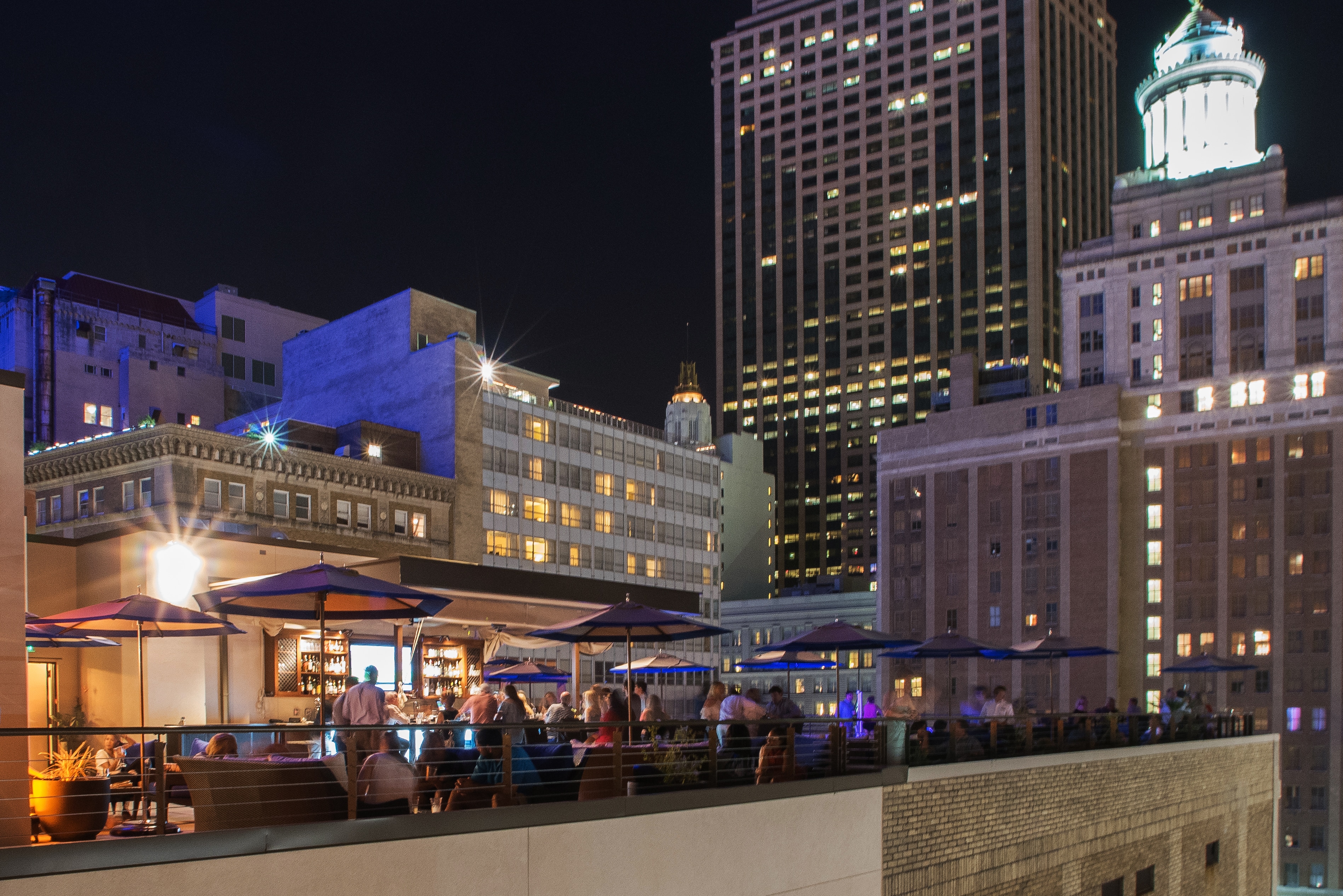 A rooftop bar with people sitting at tables under umbrellas, overlooking a city skyline at night.