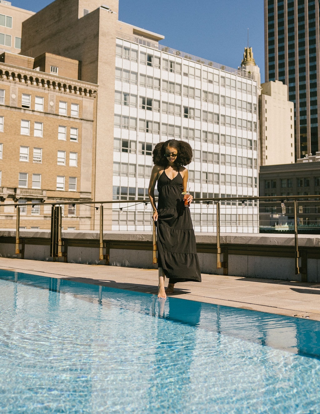 A woman in a black dress standing by a rooftop pool with a city skyline in the background.