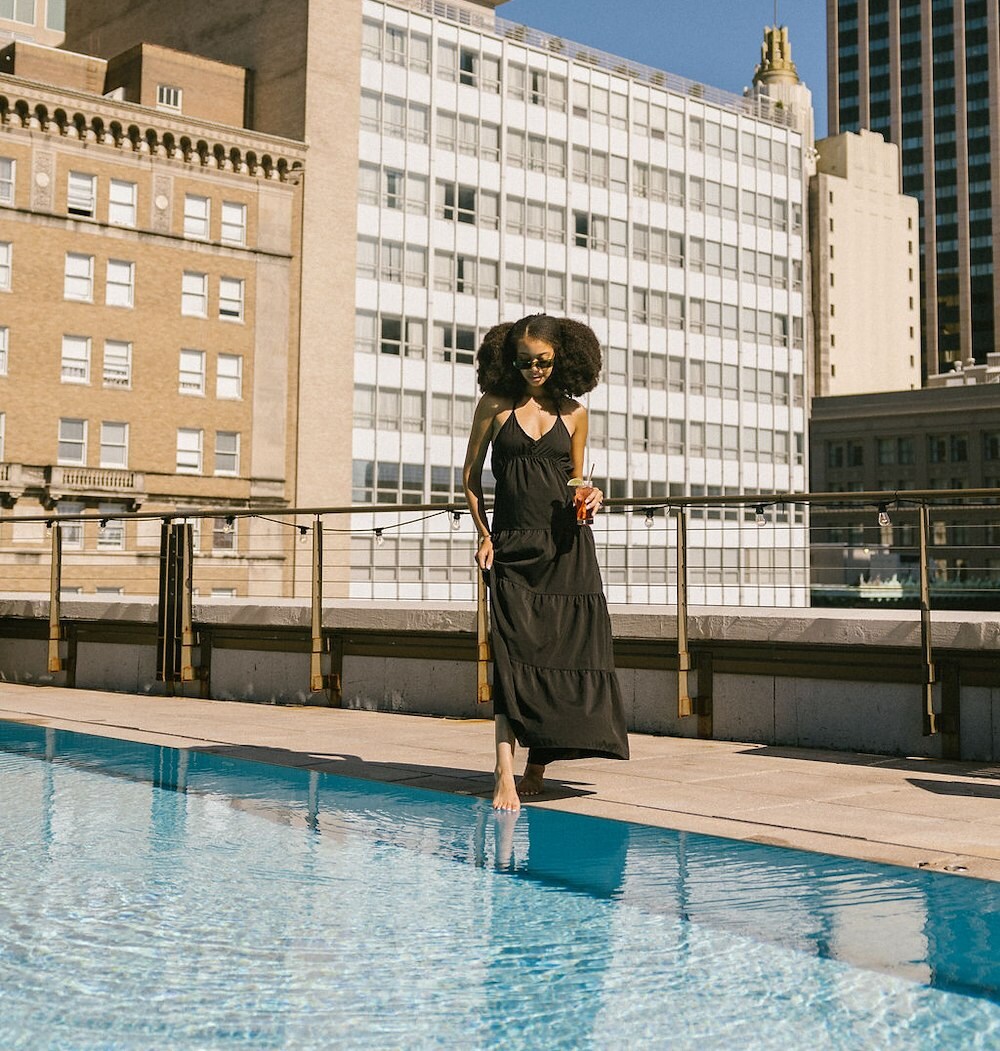 A woman in a black dress walks barefoot beside a rooftop pool, holding a drink