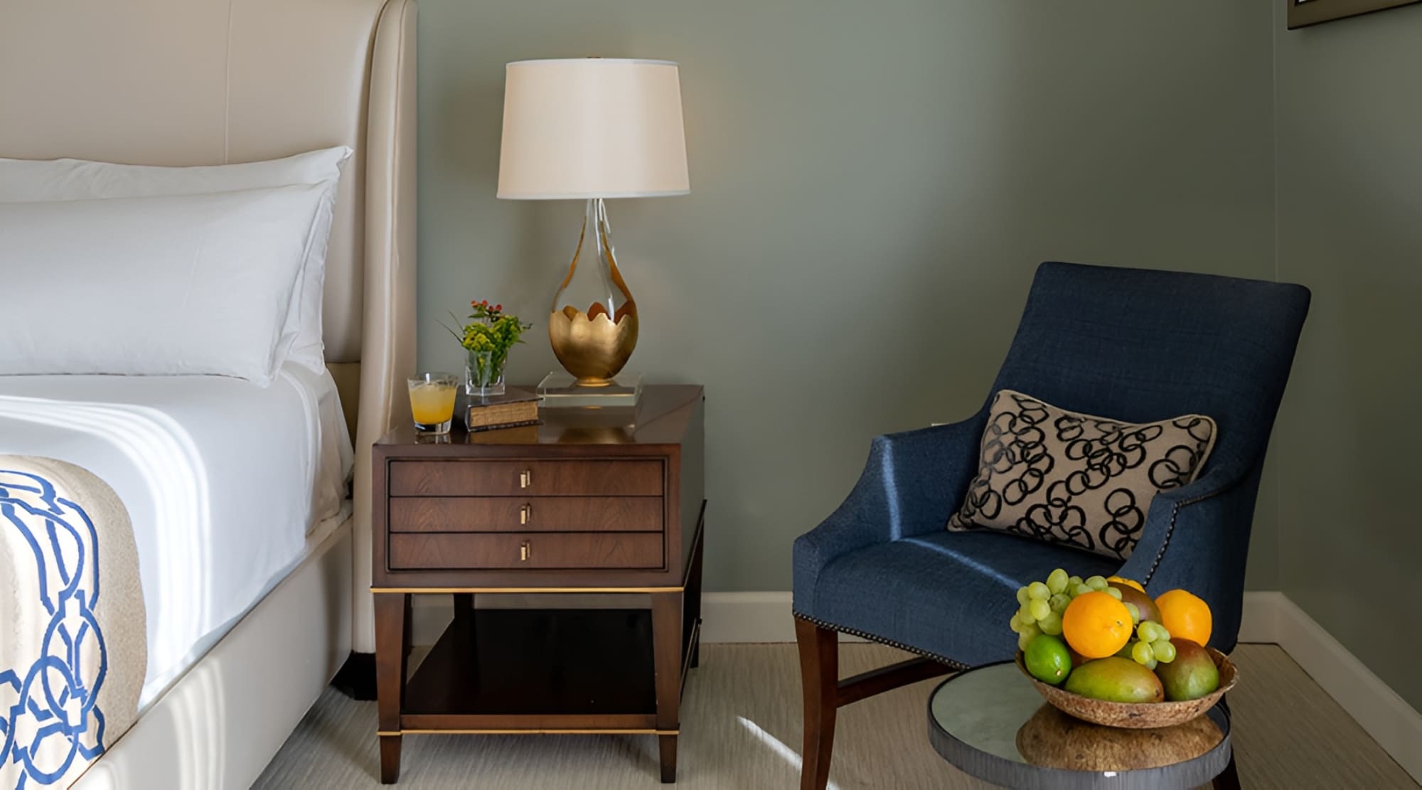 A neatly arranged hotel room with a blue armchair, a wooden nightstand, and a bed.