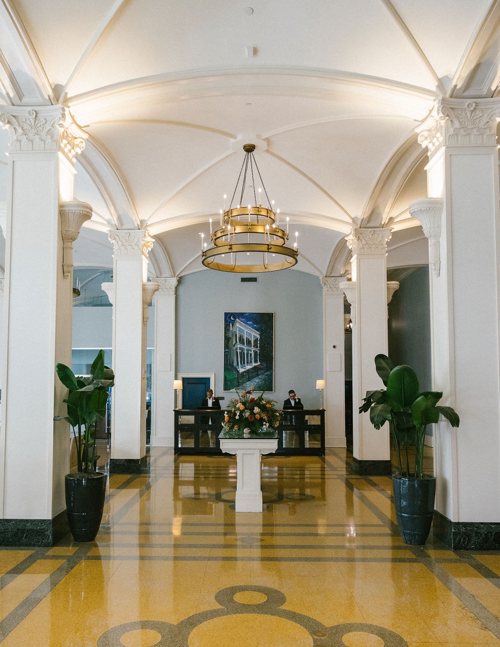 A wide view of a hotel lobby with a high ceiling, arches, columns, a chandelier, and a reception desk.
