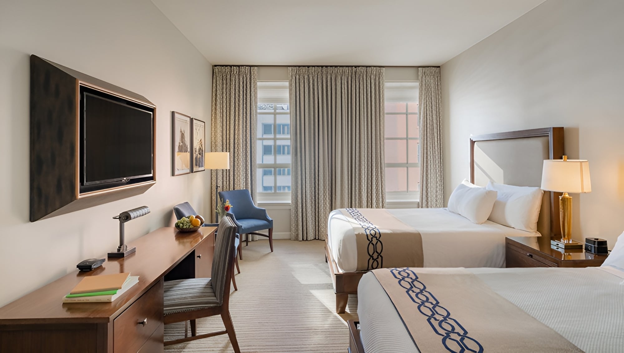 A wide view of a hotel room with two twin beds, a wood nightstand, and a gold lamp, with city view through the window.