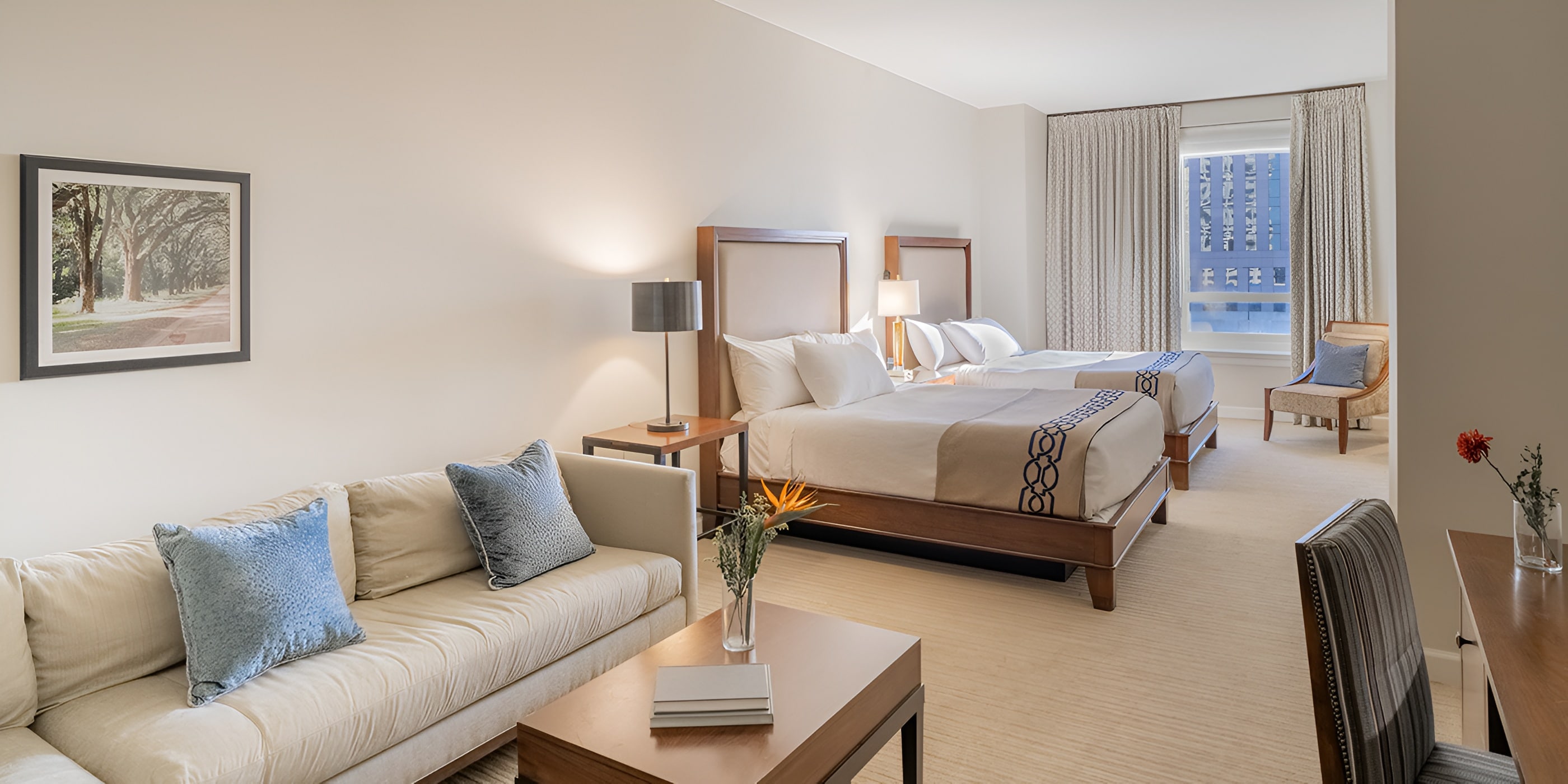 A hotel room with two beds, a window with city view, a chair, and a table with a vase of flowers.