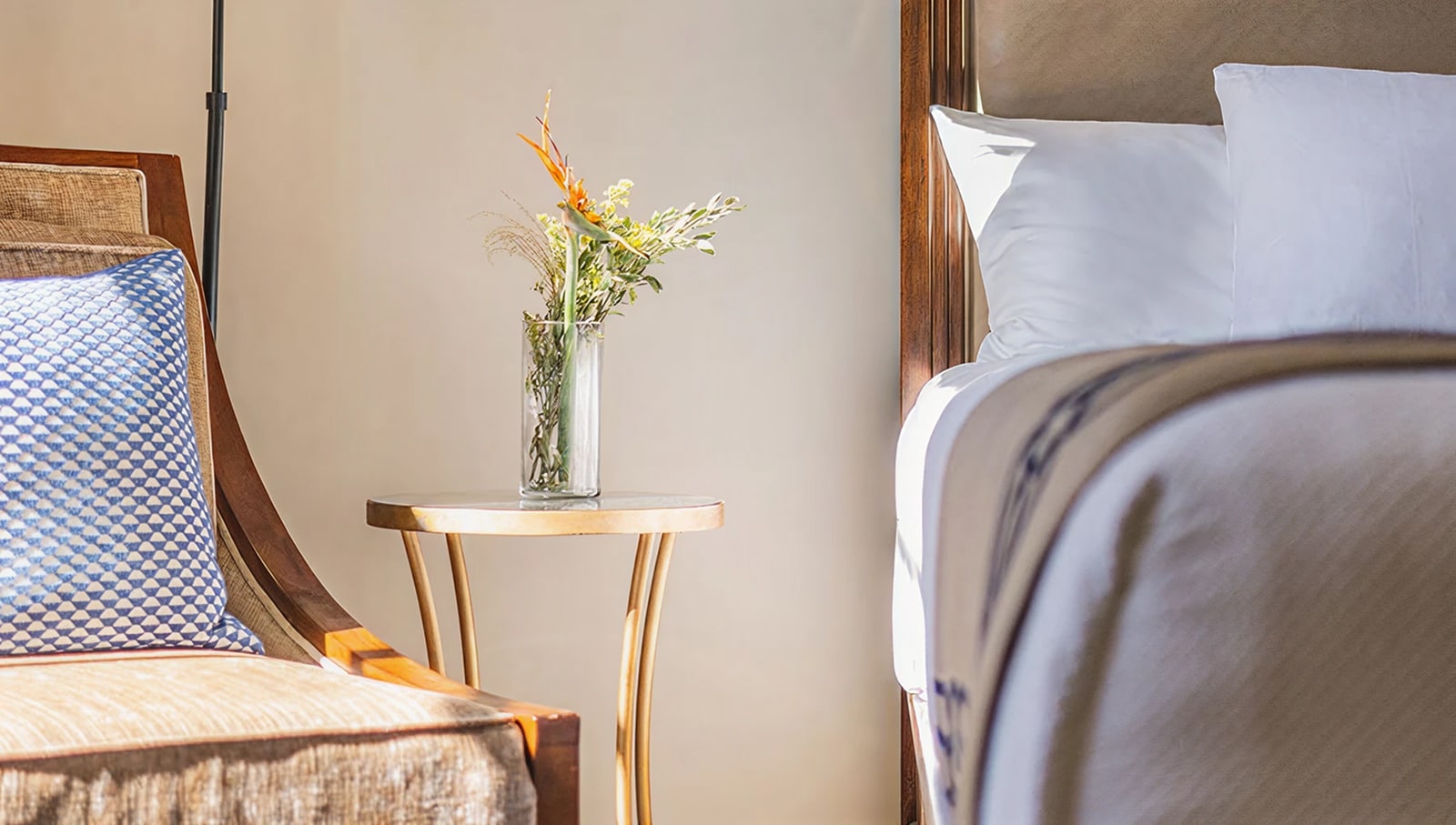 A neatly made bed with white linens and a blue and white patterned chair beside a small table with flowers.