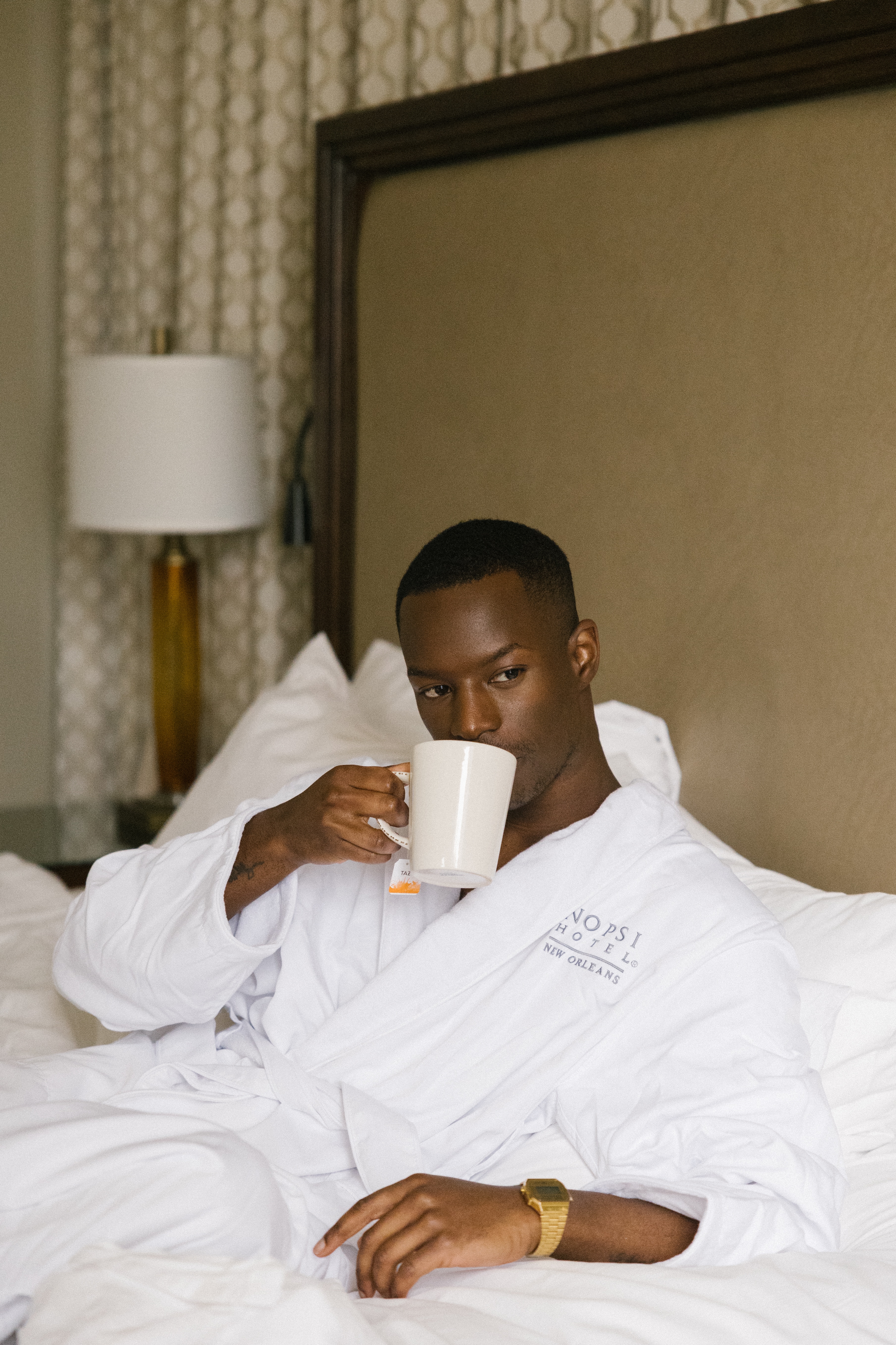 A man in a white robe sitting on a bed with a cup of coffee