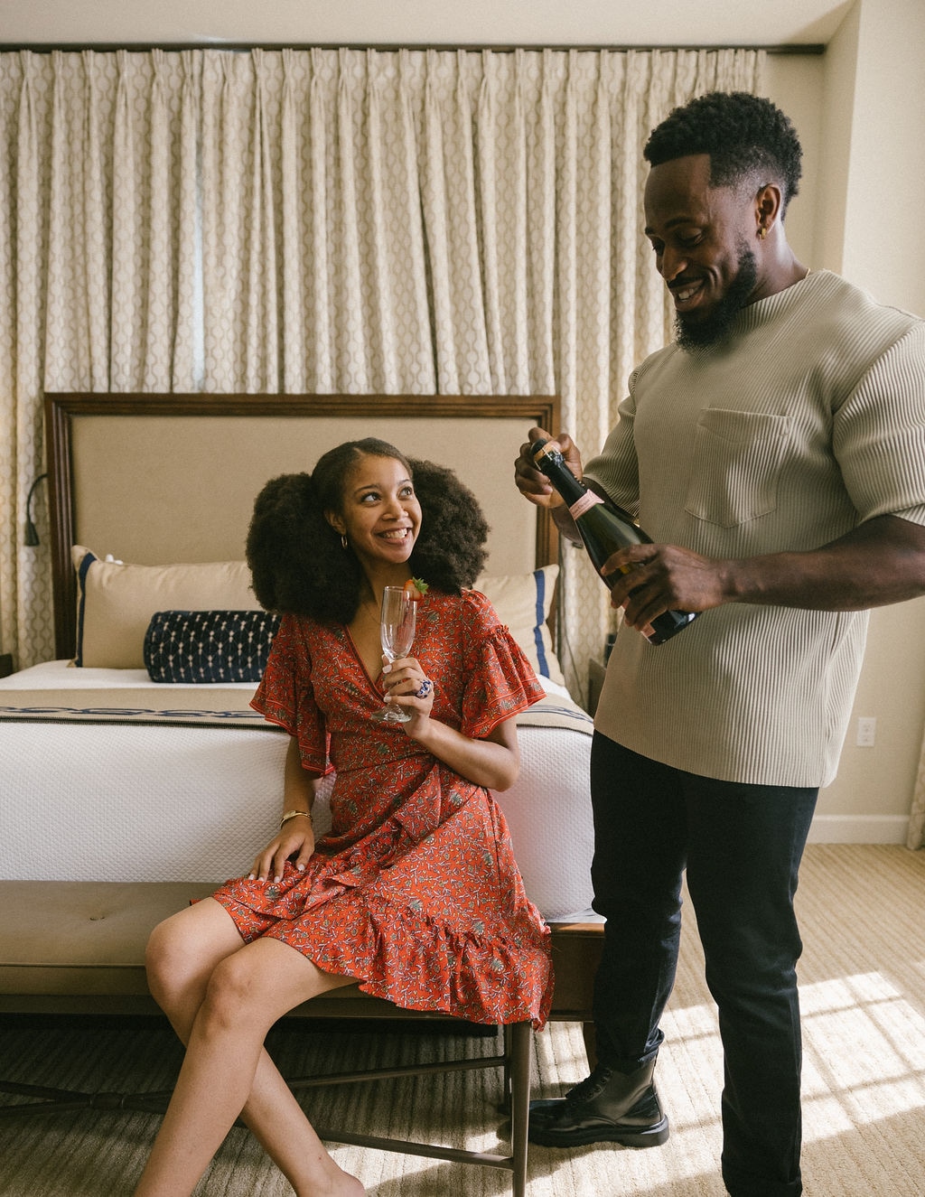 A man and woman in a hotel room with a bed, chair, and window.