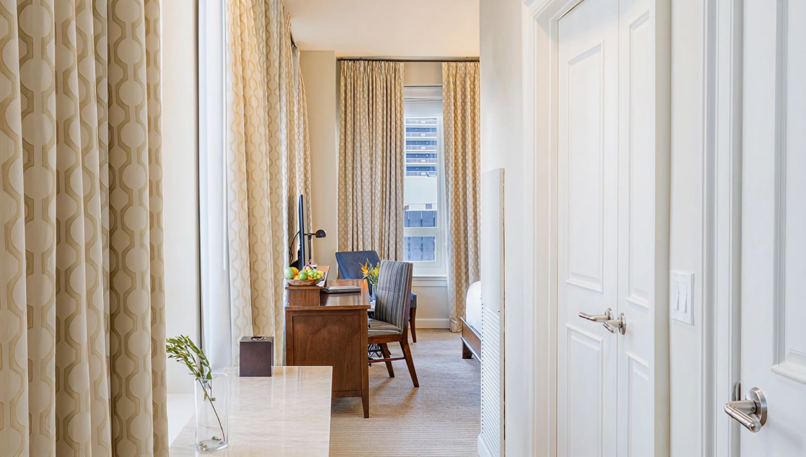 A wide view of a hotel room with a desk, chair, TV, and window