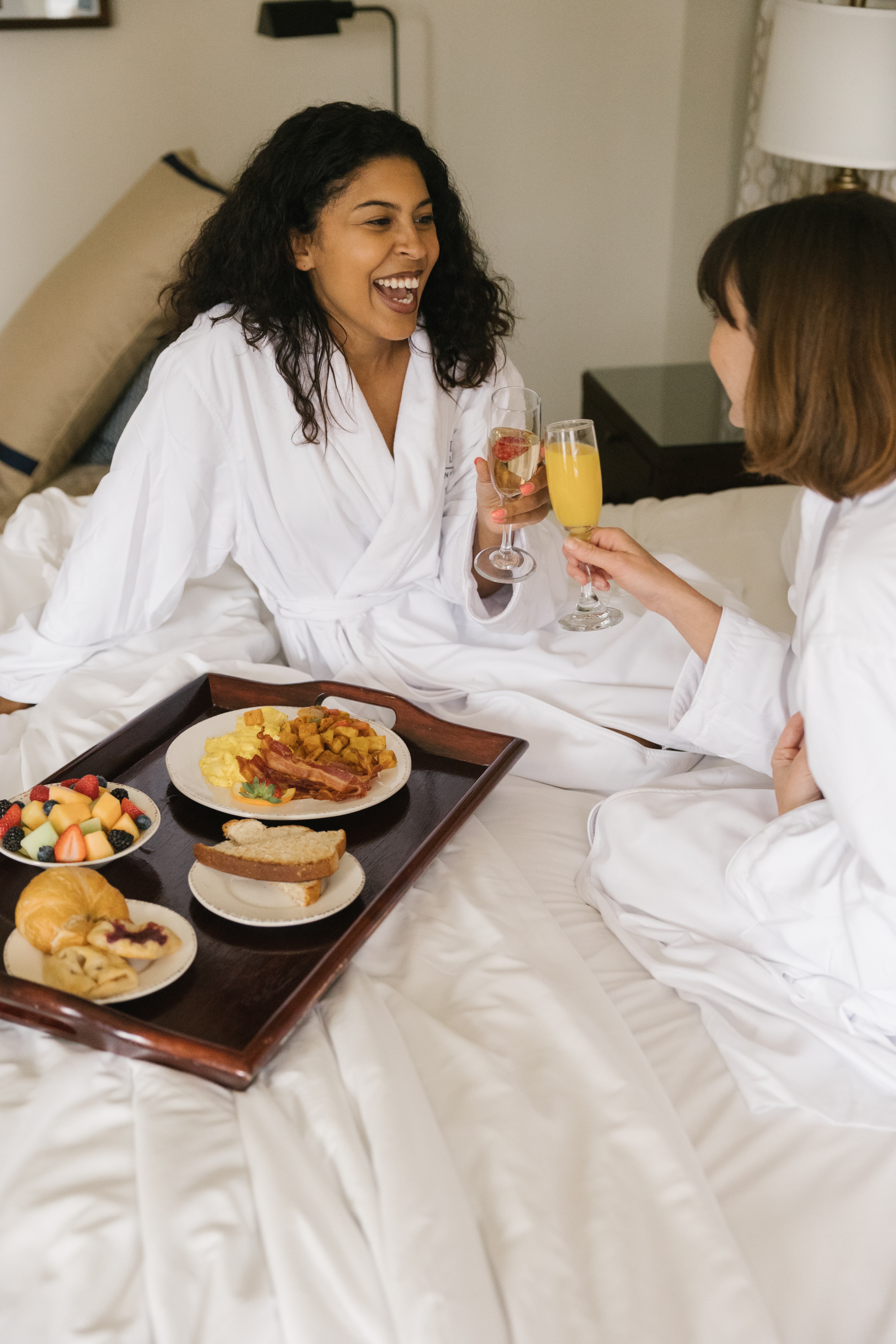 Two women in white robes sitting on a bed with a breakfast tray and champagne glasses.