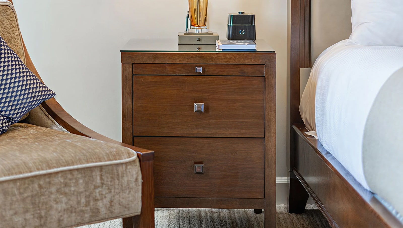 A hotel room with a wooden nightstand, a brown chair, and a bed with white linens.
