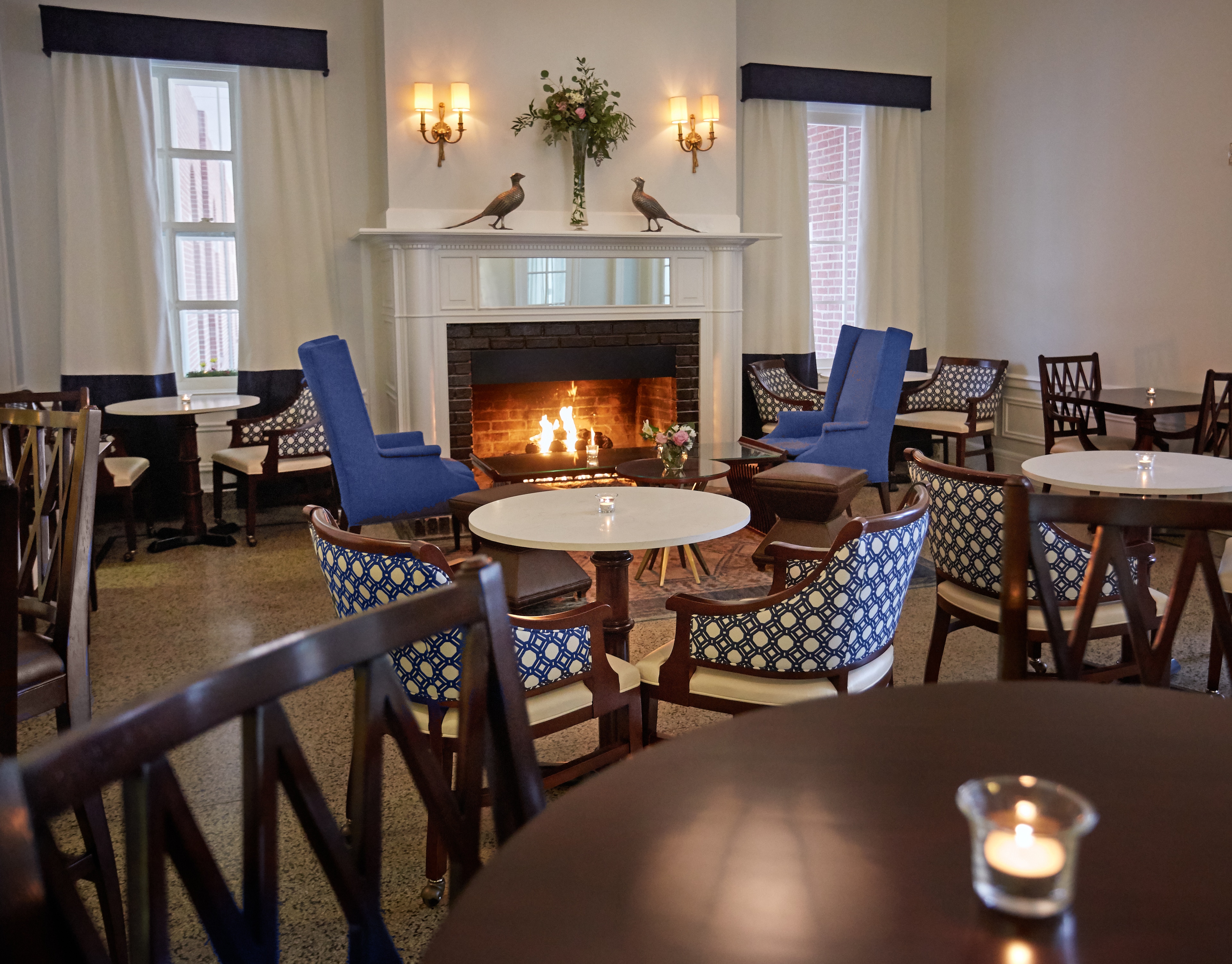 Cozy hotel lounge featuring a lit brick fireplace, blue wingback chairs, and café seating at Hotel 24 South in Staunton, Virginia