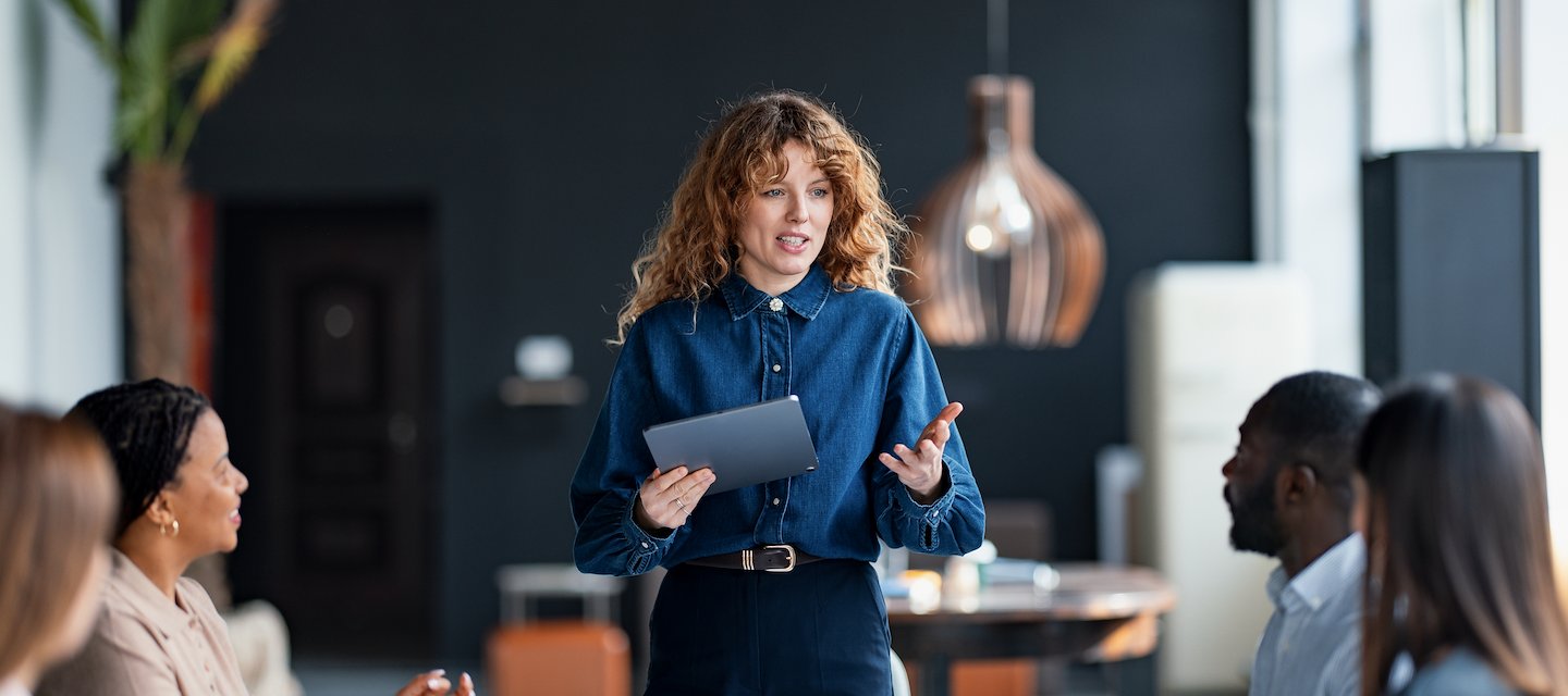 A woman in a denim shirt leads a meeting, holding a tablet, while four colleagues listen attentively around a conference table. The room is modern and well-lit.