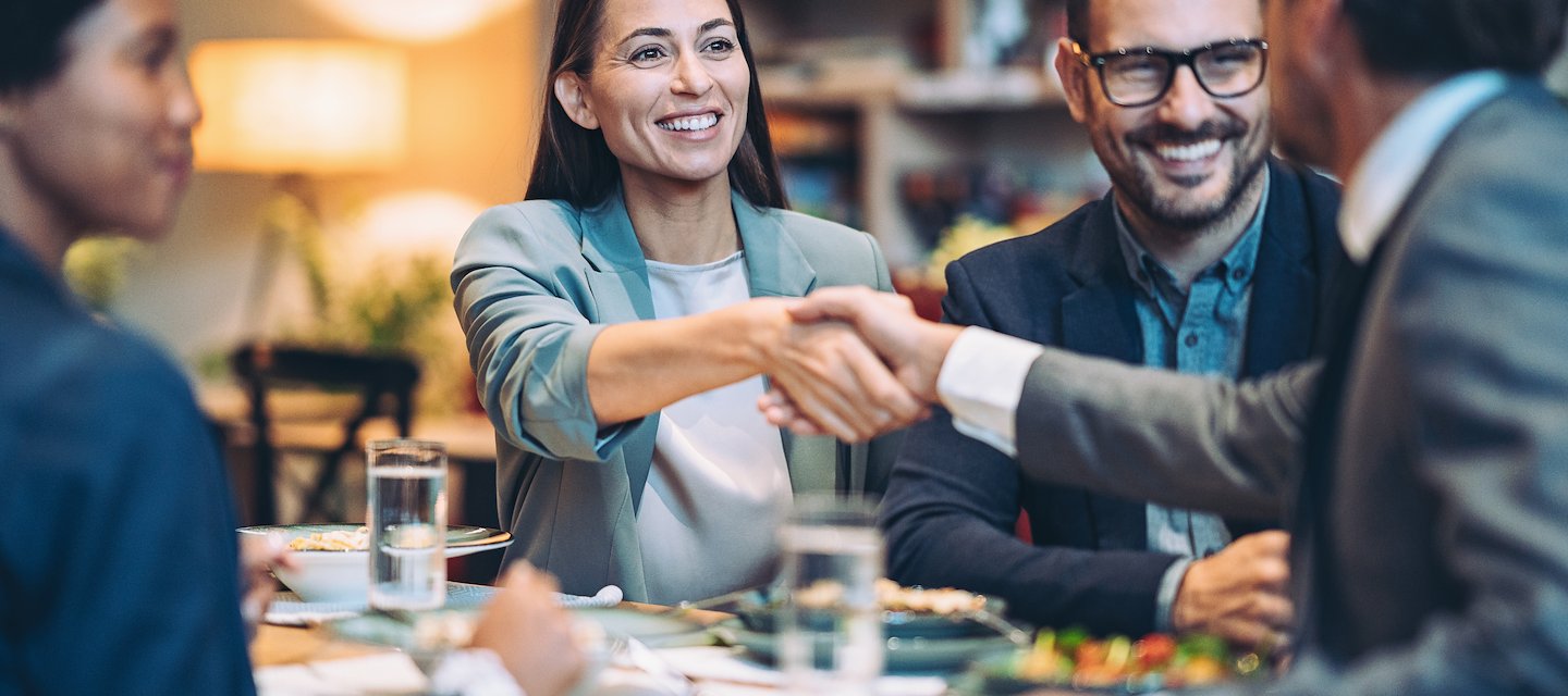 A woman smiles as she shakes hands with a man across a restaurant table, joined by two others, conveying a cheerful and collaborative mood.