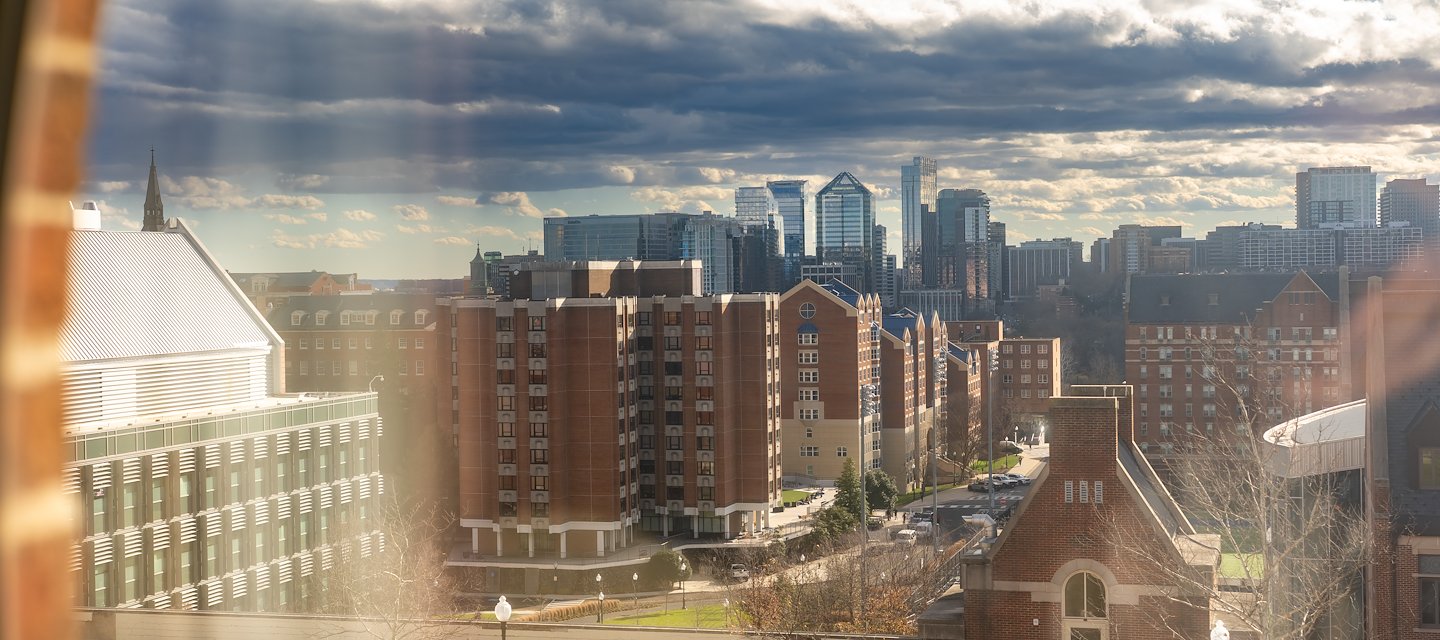 View through a window of a cityscape with brick buildings in the foreground and modern skyscrapers under a partly cloudy blue sky at The Hoya Hotel