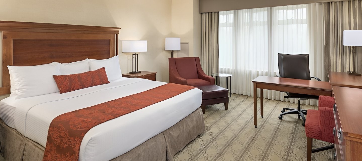 Hotel room featuring a king bed, armchair with ottoman, desk, and large window with sheer curtains at The Inn at Virginia Tech.