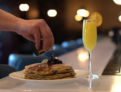 Blueberry pancakes being drizzled with warm maple syrup, alongside a mimosa.