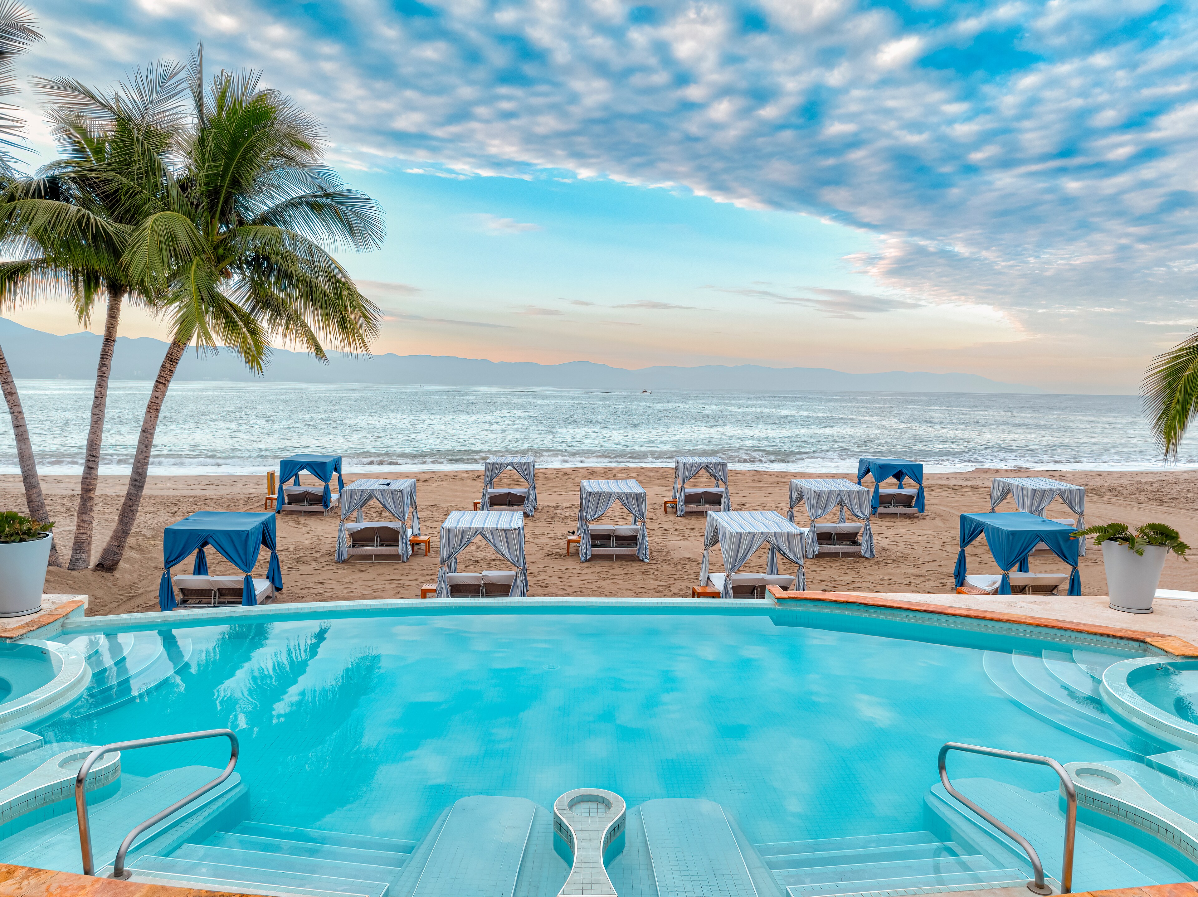 Pool overlooking beach cabanas at Casa Velas, Puerto Vallarta, with palm trees and ocean view under a blue sky.