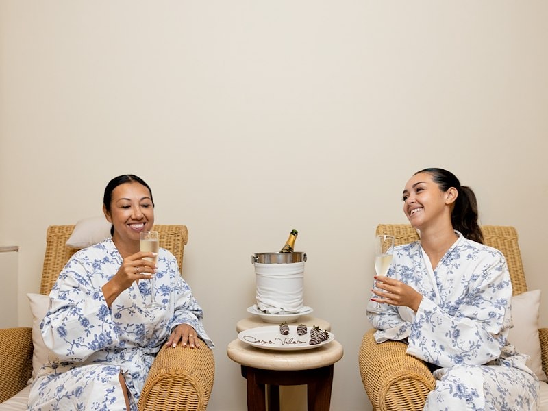 Two women in white robes sitting in wicker chairs holding glasses of champagne