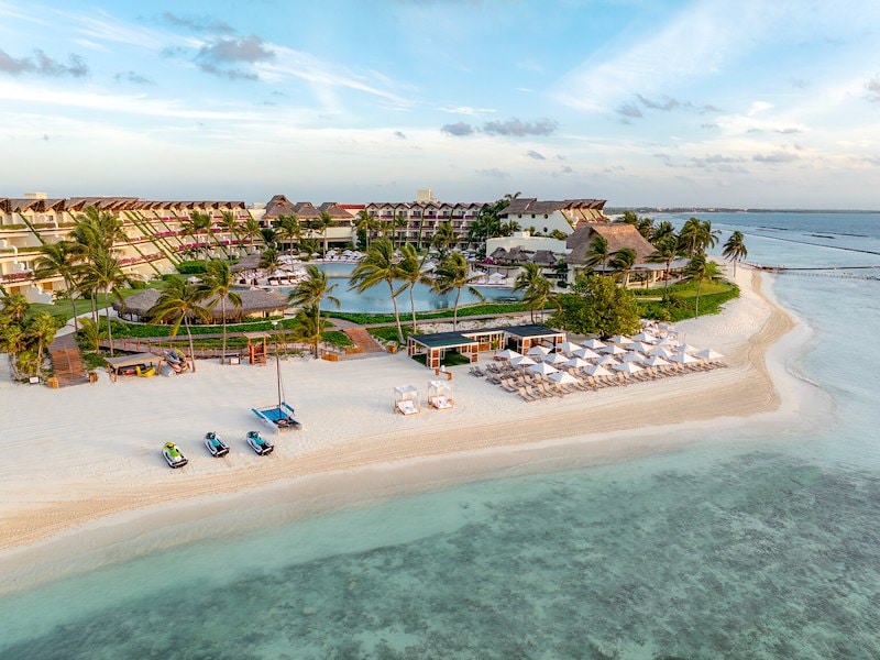 Aerial panoramic view of the Grand Velas Riviera Maya resort, featuring terraced luxury buildings with thatched roofs overlooking a pristine white sand beach and turquoise Caribbean waters. The scene includes several jet skis and a catamaran on the shore, rows of white umbrellas with lounge chairs, lush palm trees, and a large beachfront swimming pool under a soft blue sky.