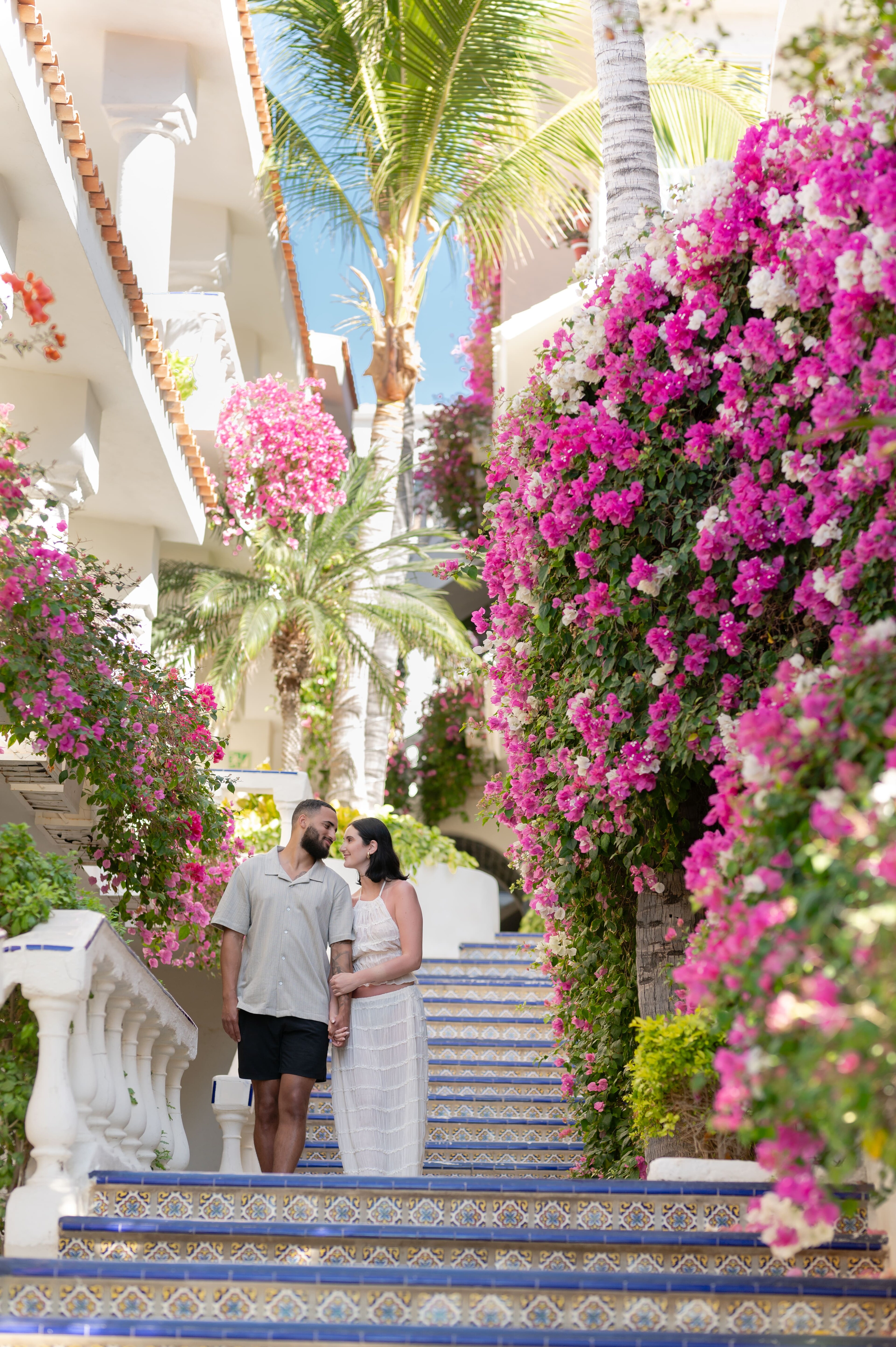 Couple holding hands on colorful tiled steps, surrounded by vibrant bougainvillea at Mar del Cabo