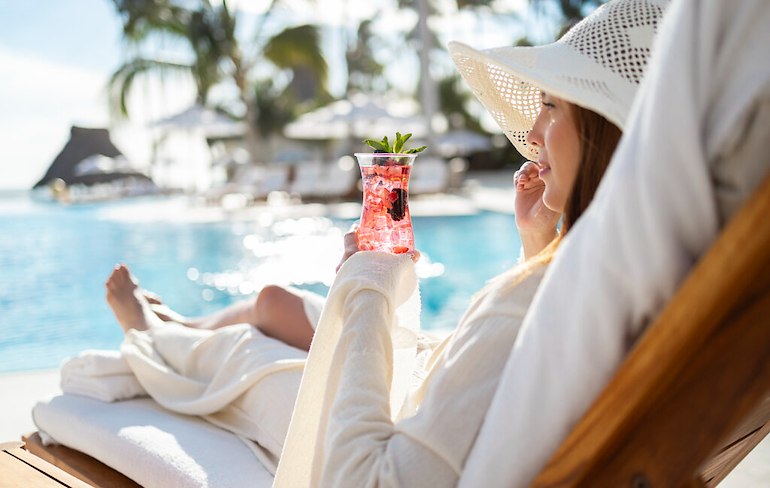 A woman in a white hat holding a drink while lying on a lounge chair