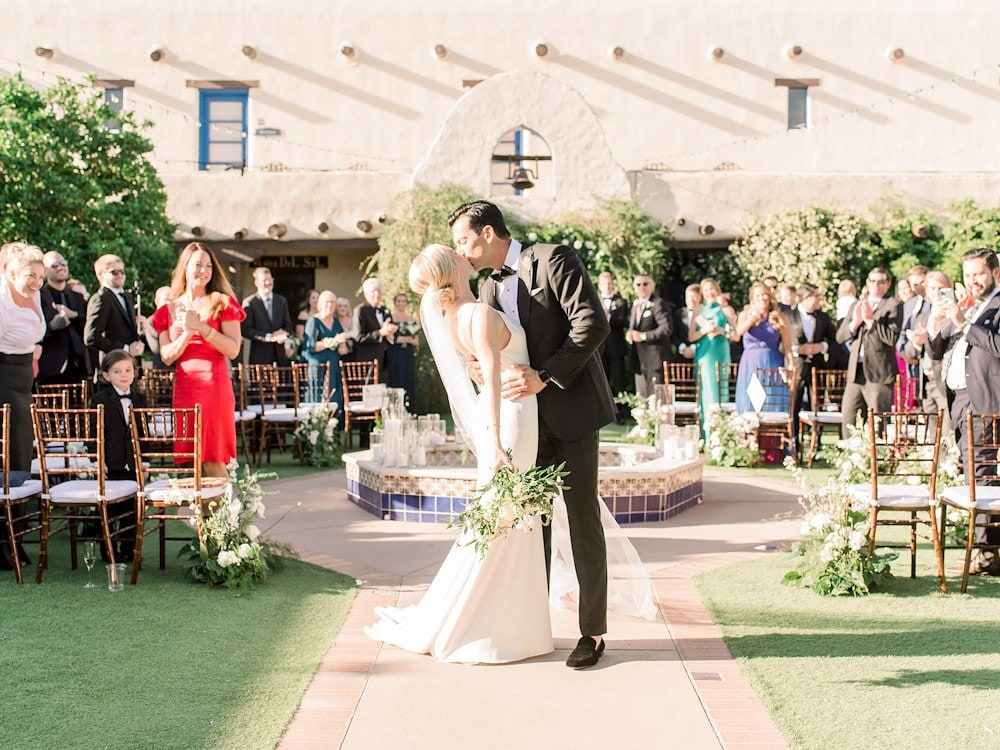 A newlywed couple shares a kiss in a sunlit garden courtyard
