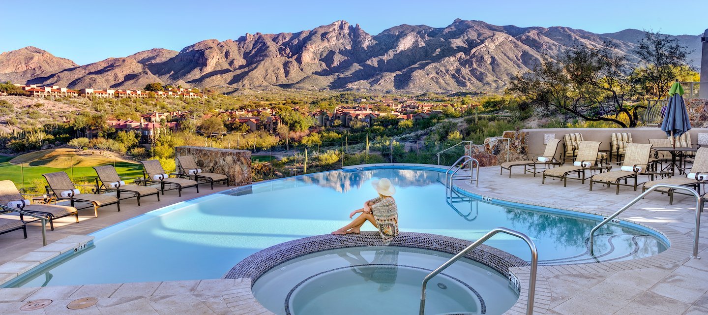 Una persona con un sombrero de sol se relaja al lado de una piscina con vista a una pintoresca montaña