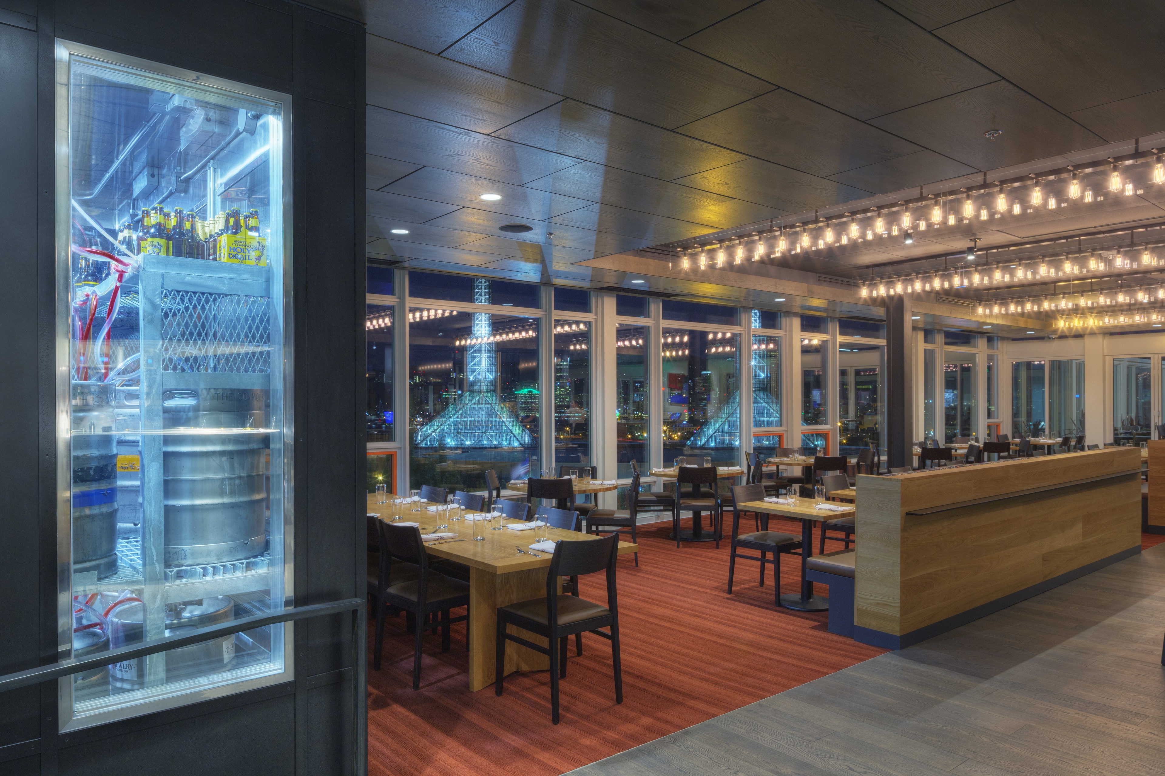 A dining area with tables and chairs, a bar, and large windows showing a city view at night.