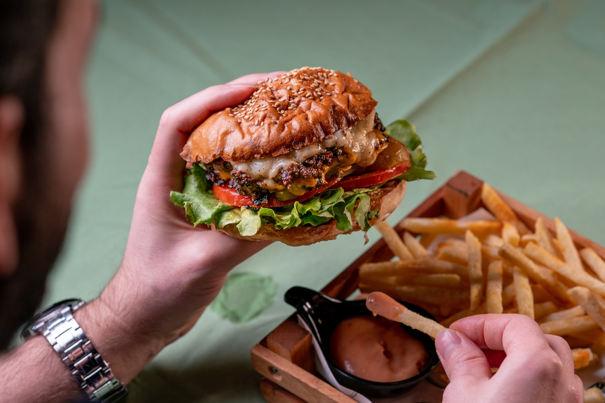 a hand holding a burger over a table with fries and sauce