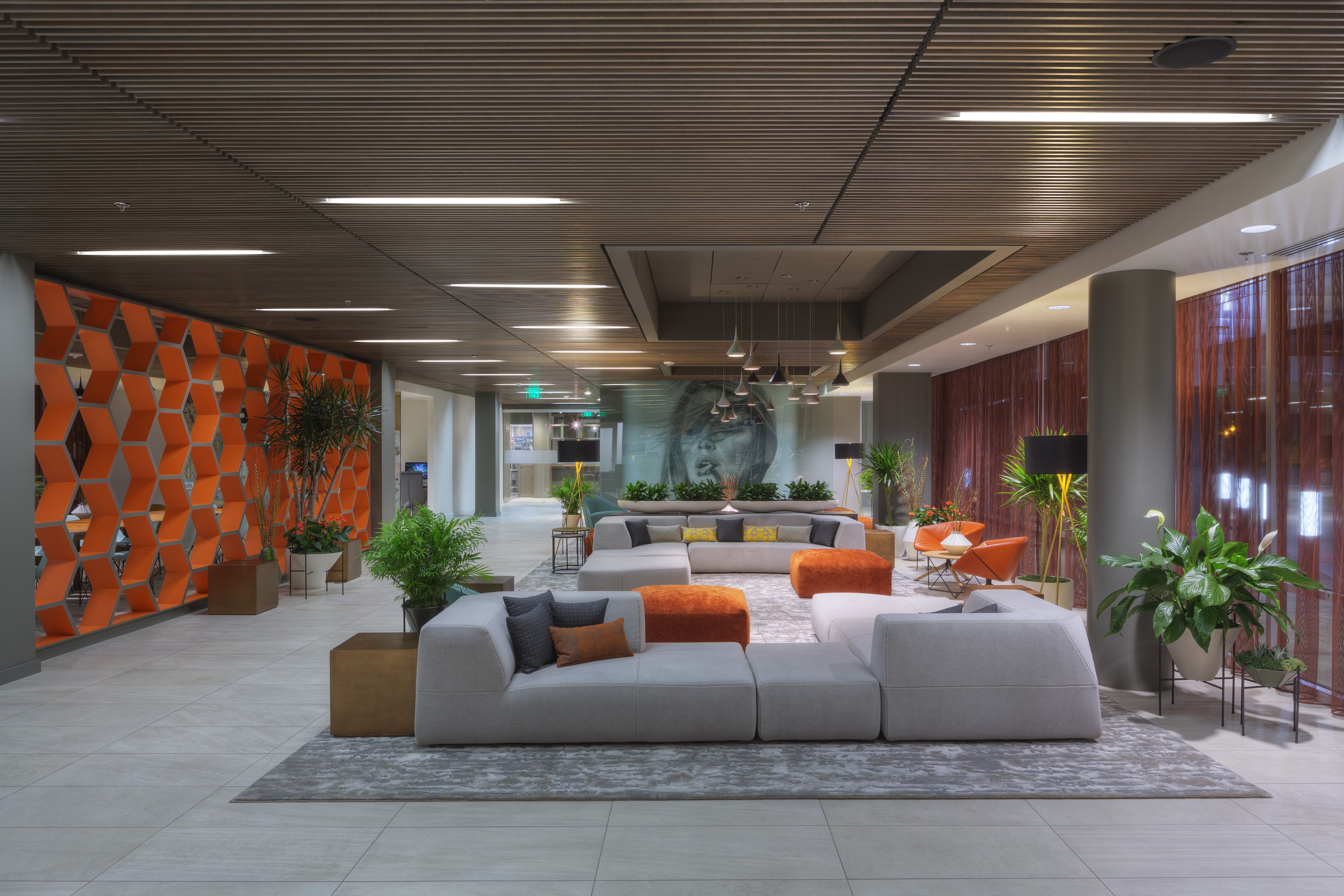 A modern hotel lobby with a gray sectional sofa, orange accent pillows, and a large plant-filled area.