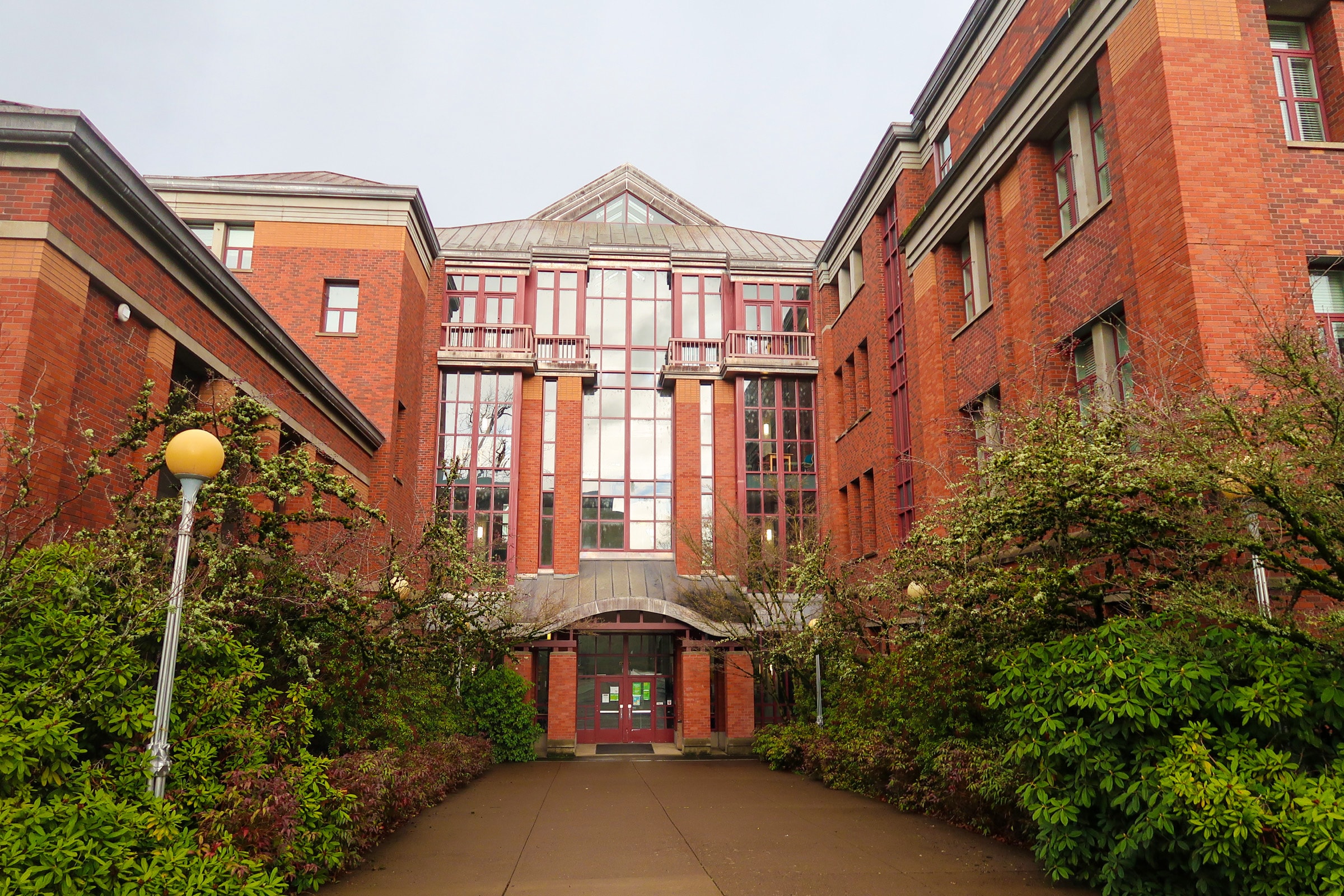 A red brick building with glass windows and balconies, surrounded by landscaping
