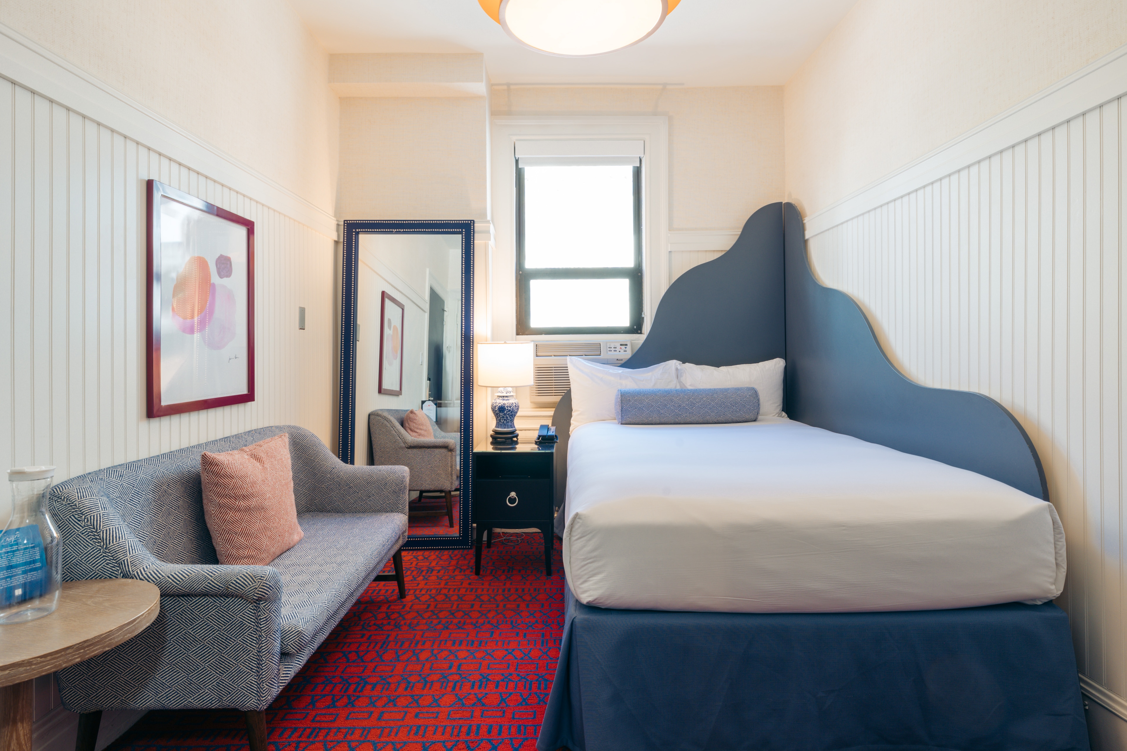 A hotel room with a bed, sofa, nightstand, mirror, and window