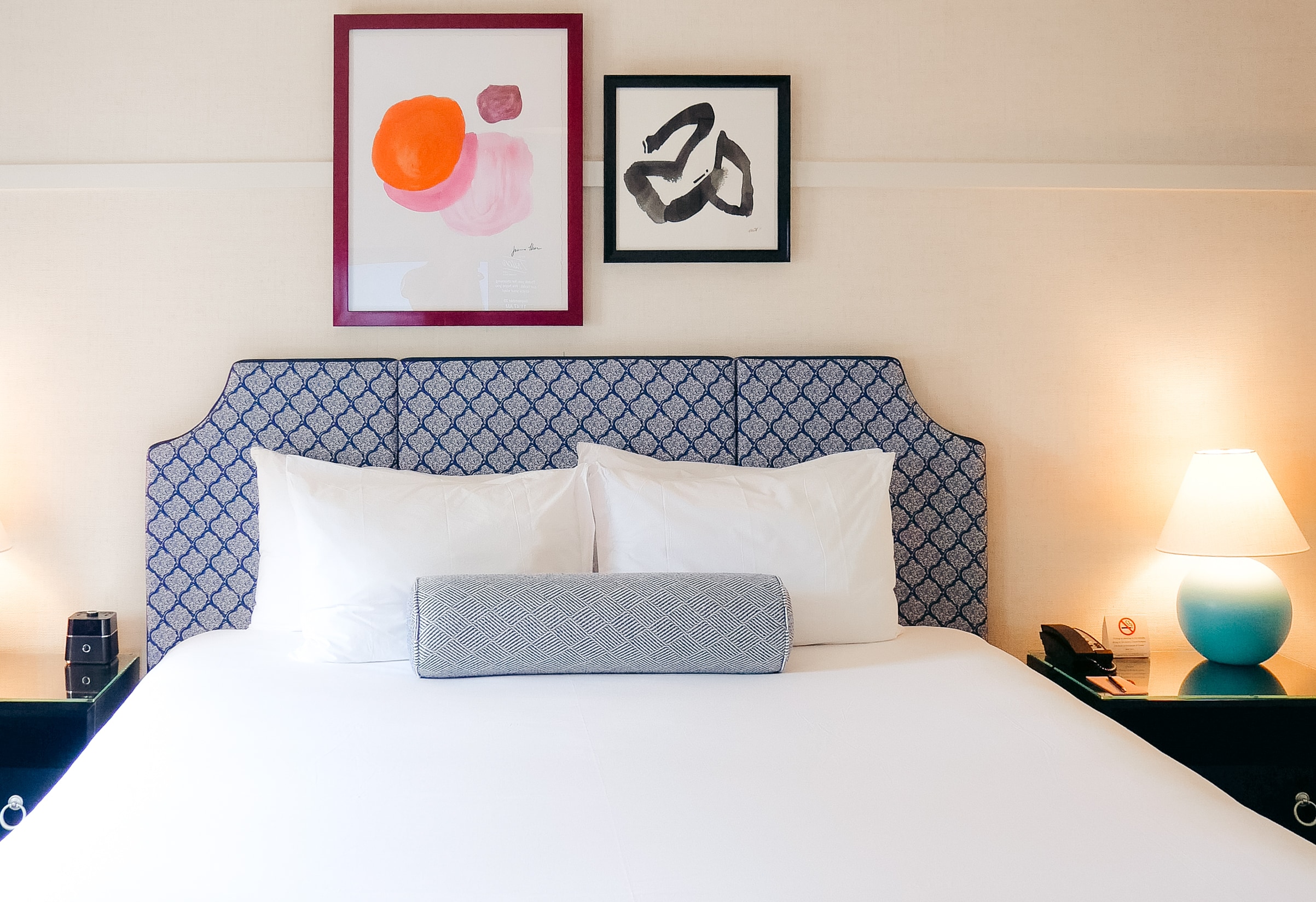 A neatly made bed with a blue and white patterned headboard, white pillows, and two pieces of abstract artwork above it.