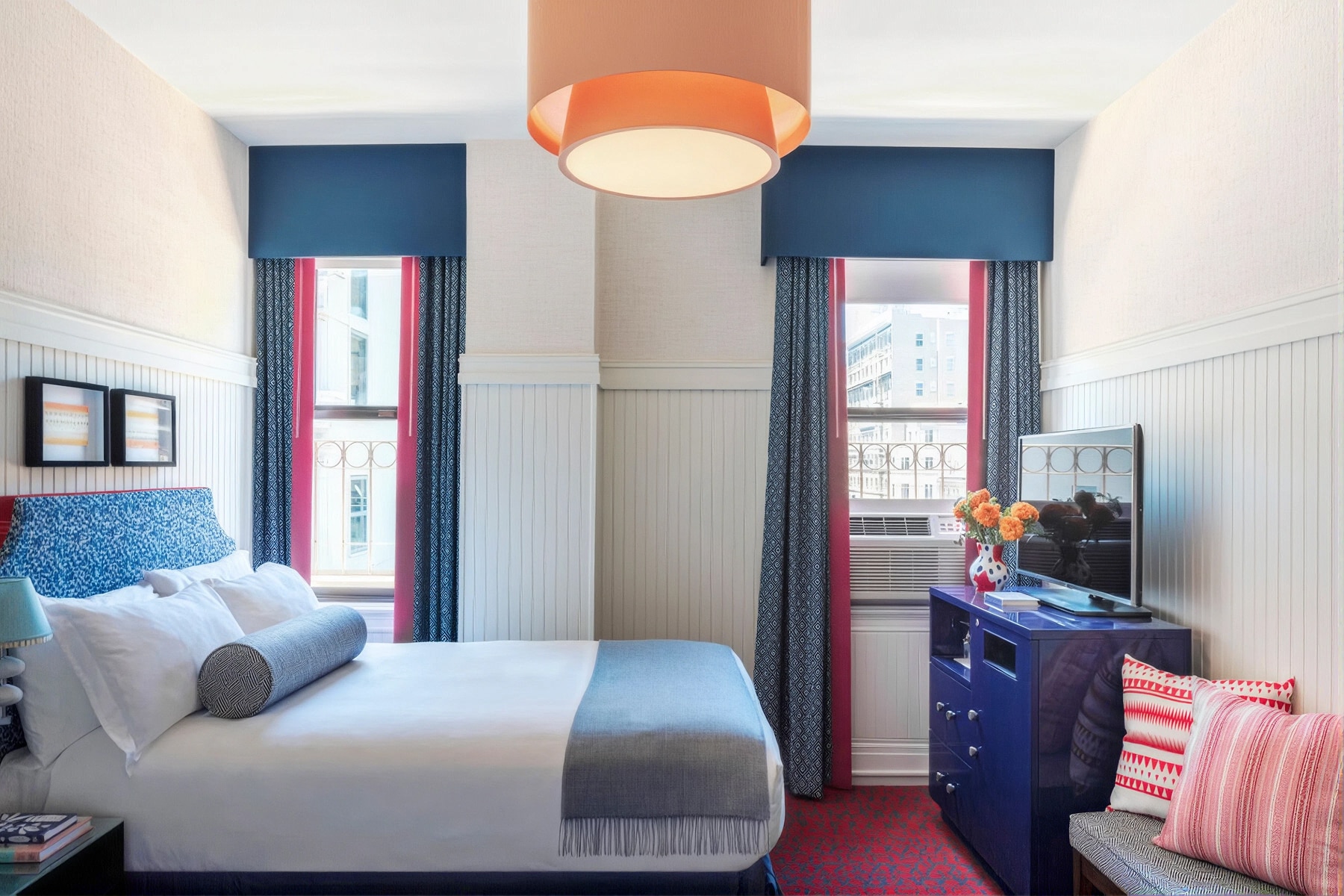 A hotel room with a bed, two windows, a dresser with flowers, and a lamp.