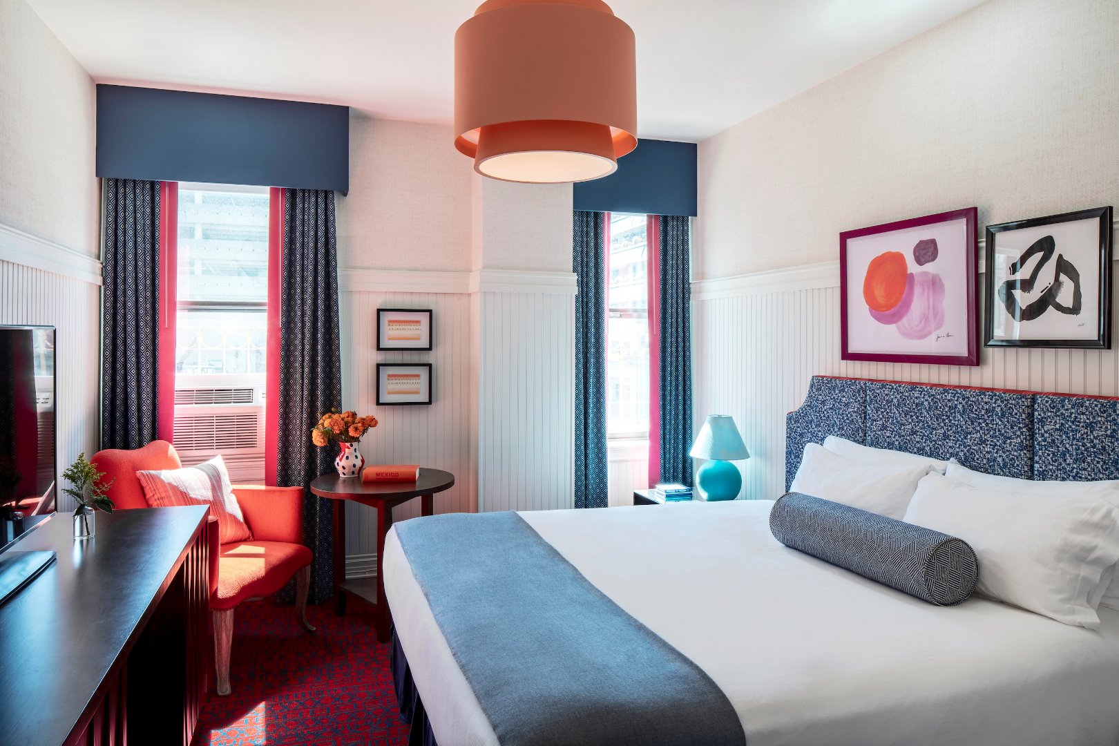 A hotel room with a bed, desk, chair, and artwork on the walls.