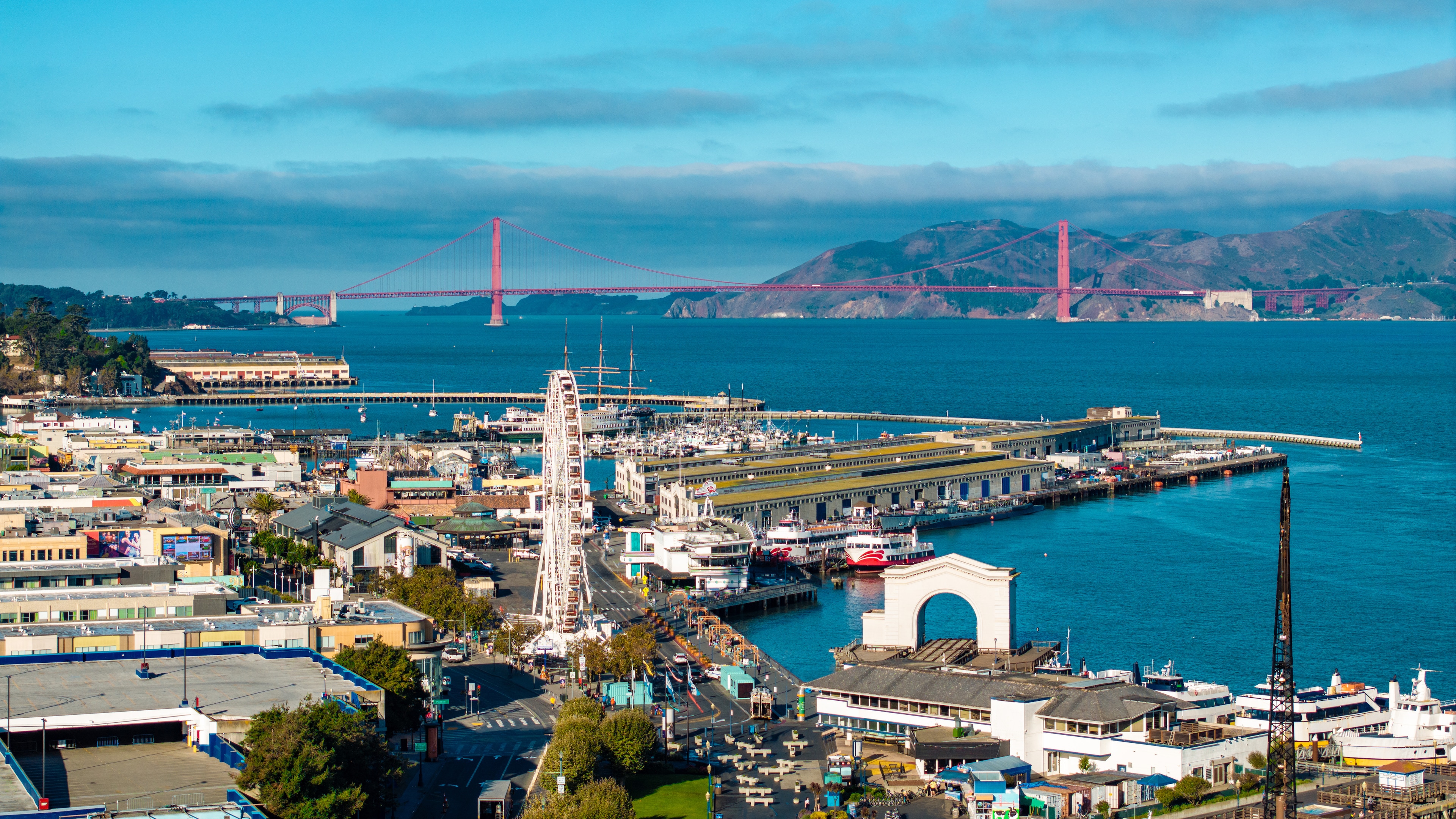 A wide-angle view of a cityscape with a large body of water, a bridge, and mountains in the background.