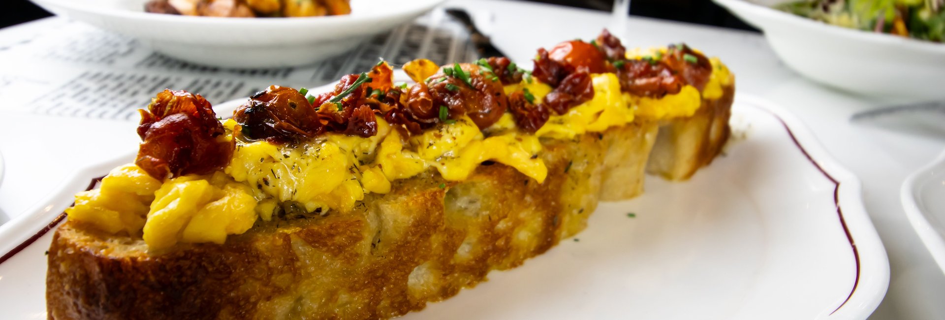 A long piece of crispy bread topped with scrambled eggs, roasted cherry tomatoes, and herbs on a white plate at Il Solito.