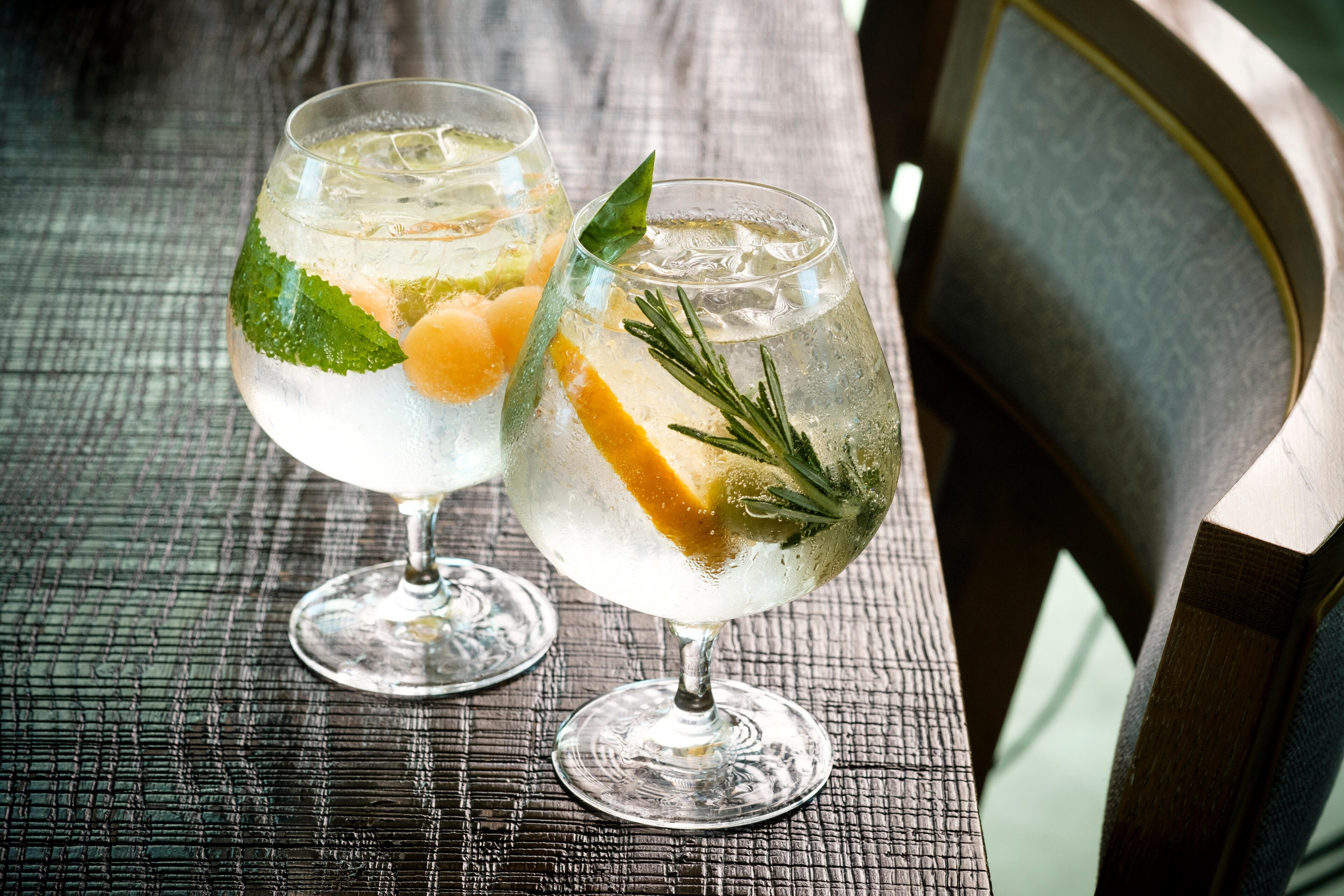 Two gin and tonics on a wooden table, one infused with melon balls and mint, the other with an orange slice and rosemary, exuding a refreshing vibe at The Soleil Beach Club Florida