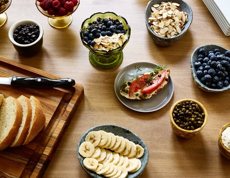 Breakfast spread with fresh bread, berries, bananas, yogurt, nuts, and toast with tomato and herbs.
