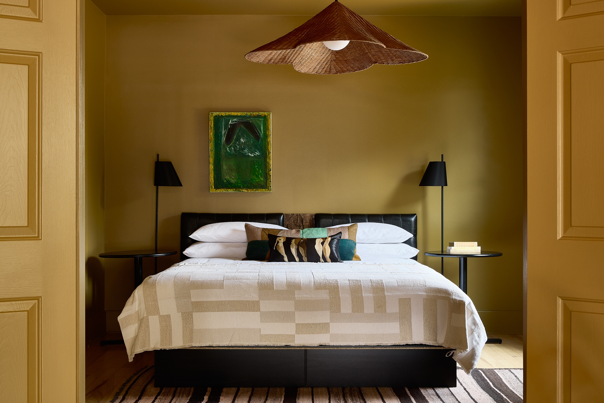 A neatly made bed with a white comforter and several pillows, flanked by two nightstands with lamps, in a room with yellow walls and a piece of green artwork above the bed.