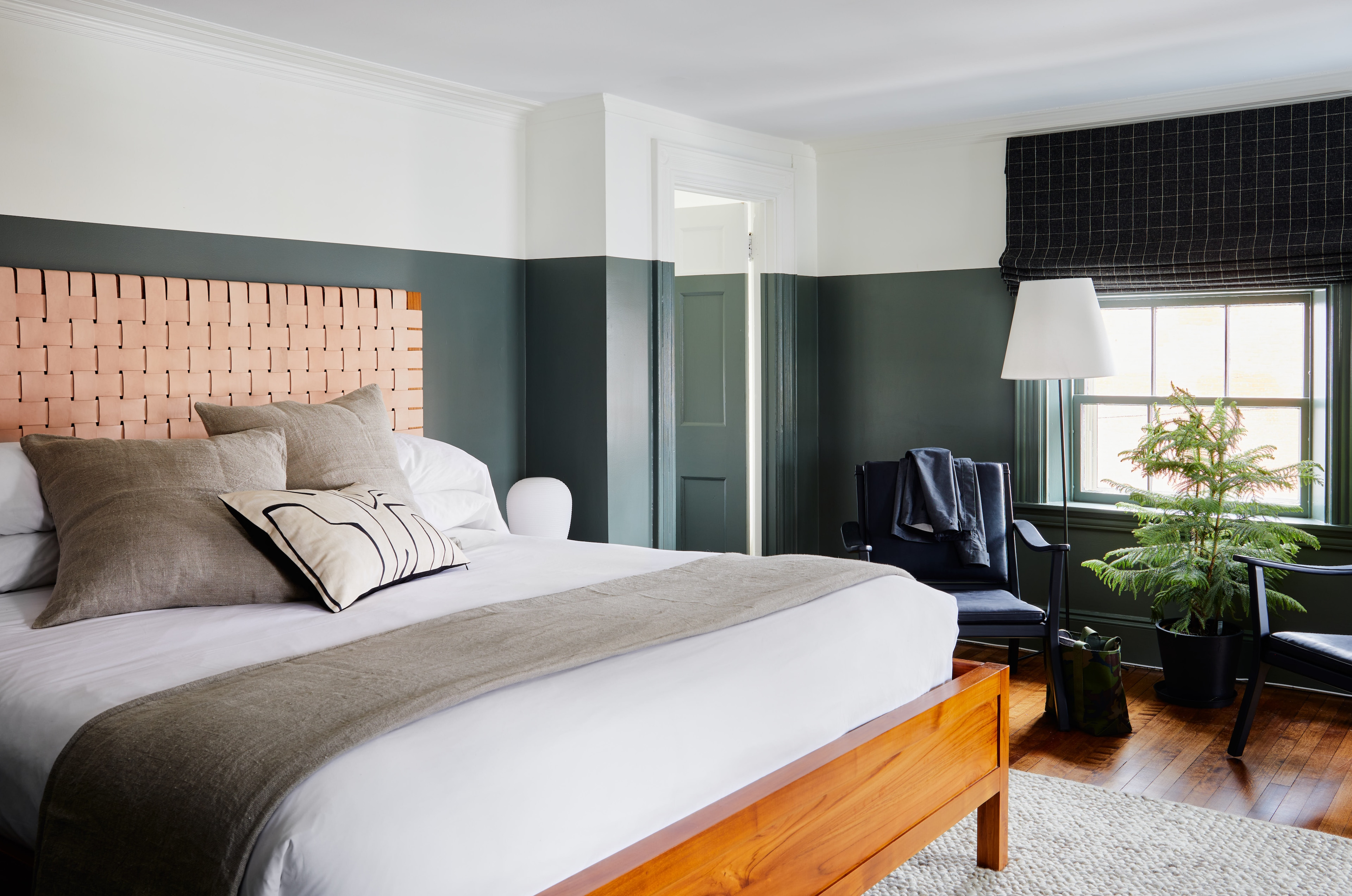A neatly made bed with pillows and a throw blanket in a room with a window, chair, and plant.