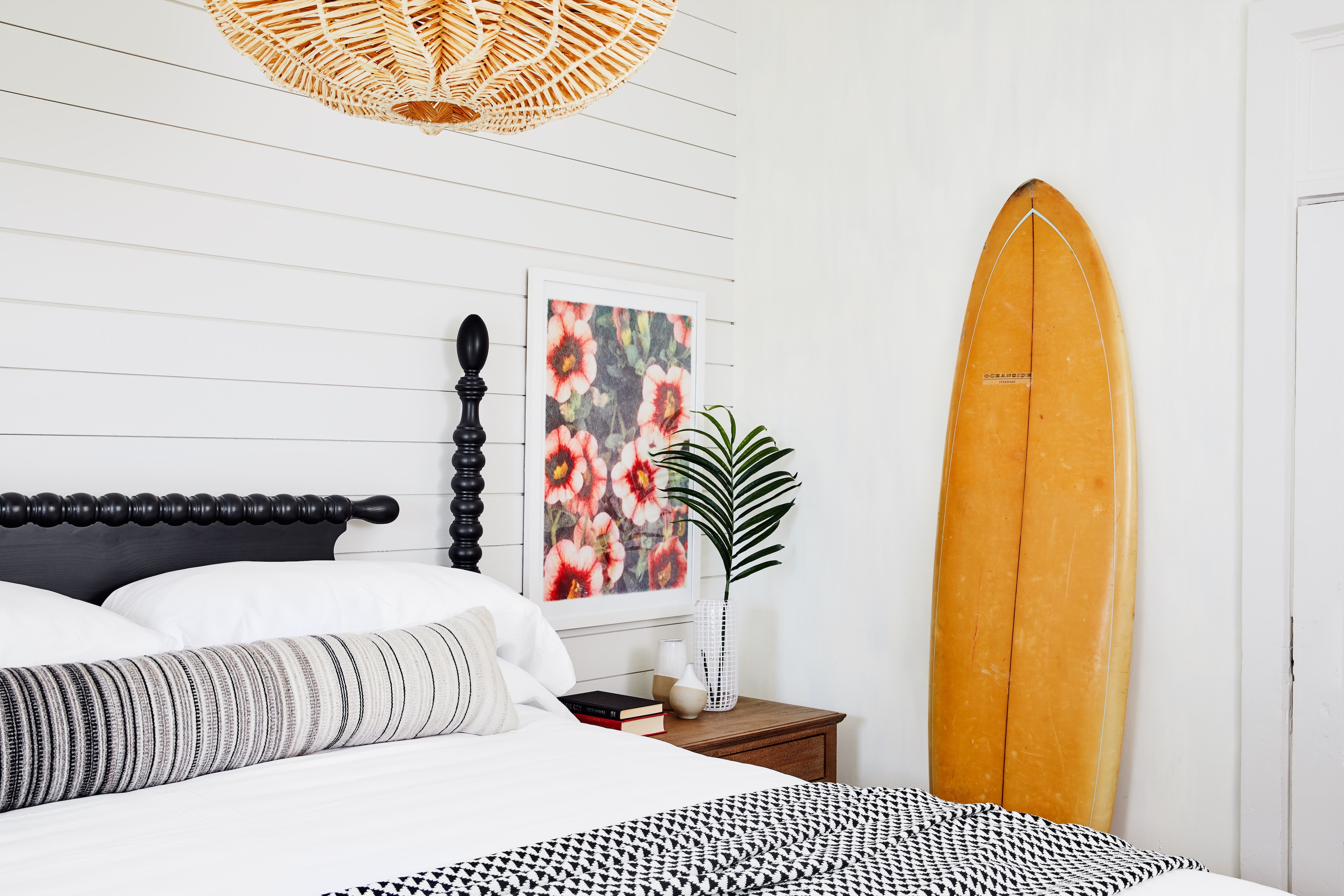 A bed with white linens and a black headboard, with a surfboard leaning against the wall next to it.